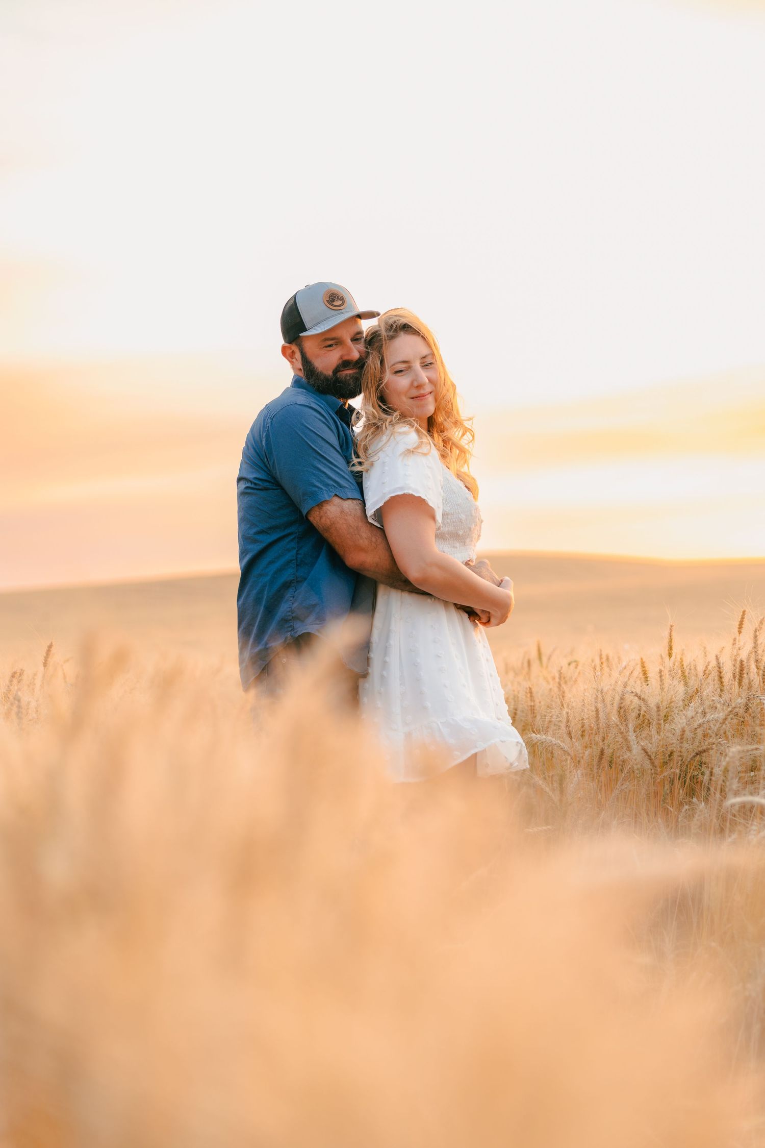 Couple embracing in a golden wheat field at sunset, man in blue shirt and cap, woman in white dress.