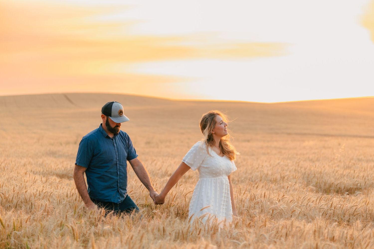 Couple holding hands in golden wheat field at sunset, woman in white dress and man in blue shirt.