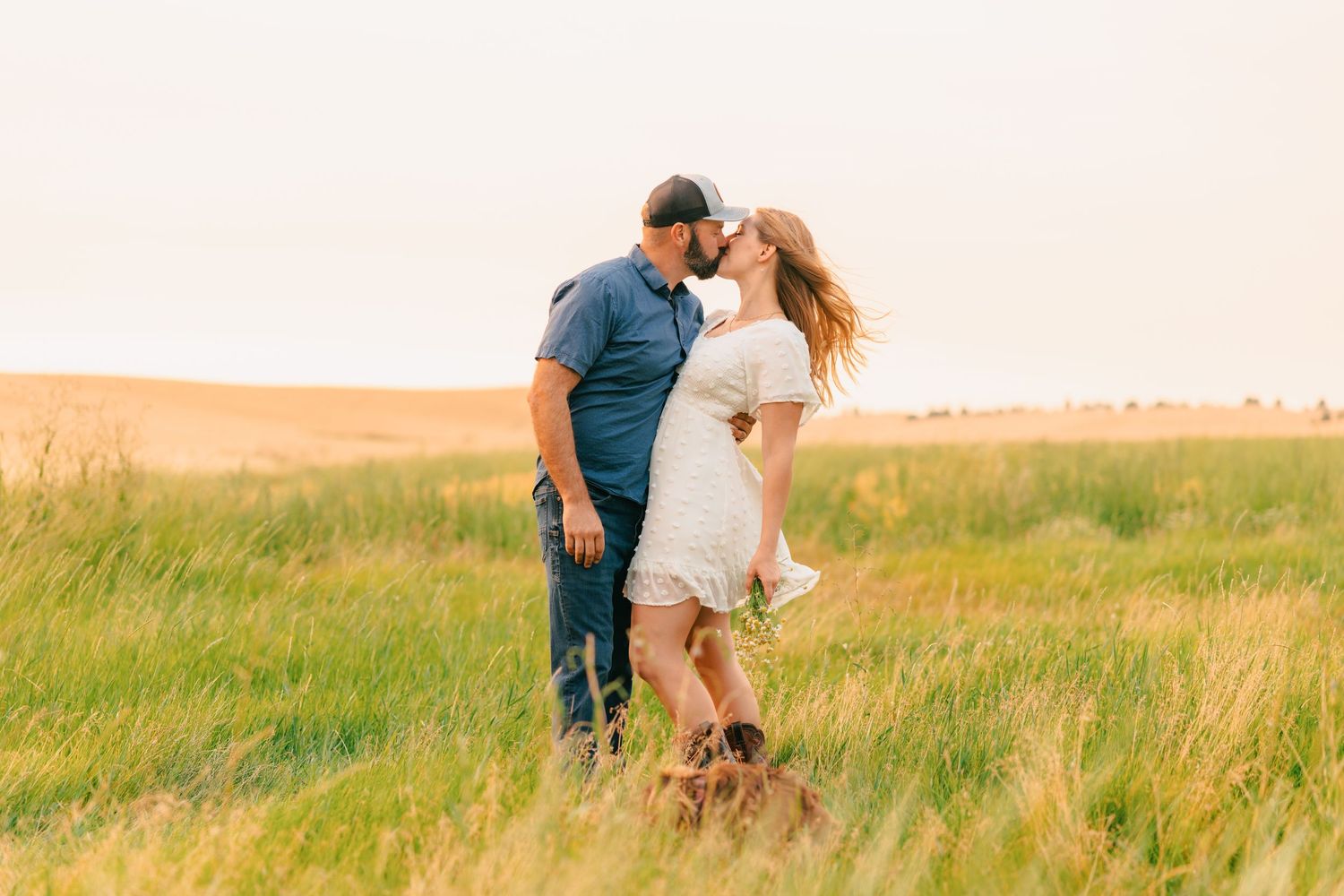 Couple sharing a romantic kiss in a lush green field at golden hour, woman in white dress, man in blue shirt.