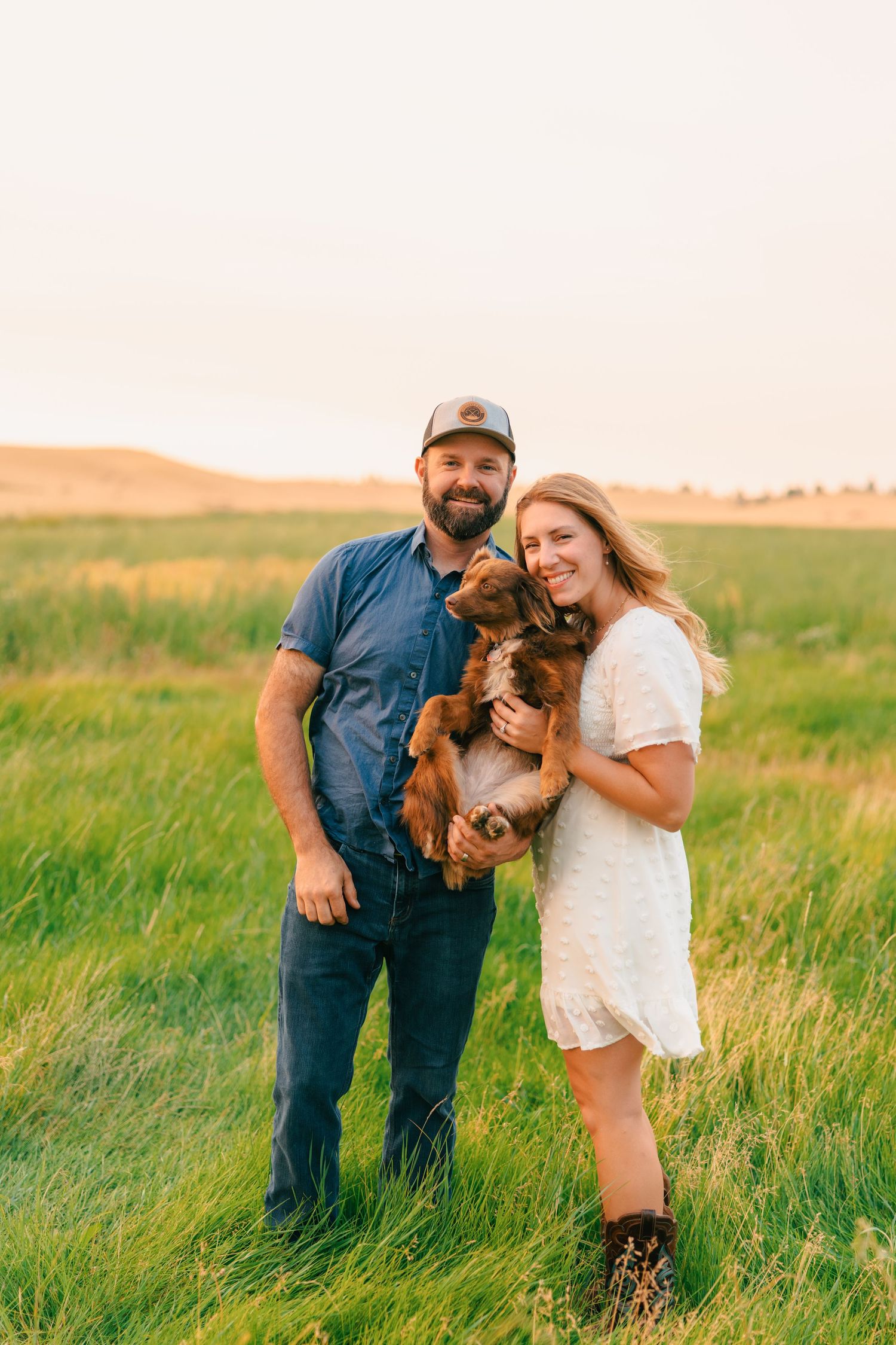A smiling couple holds a brown dog in a lush green field at golden hour.