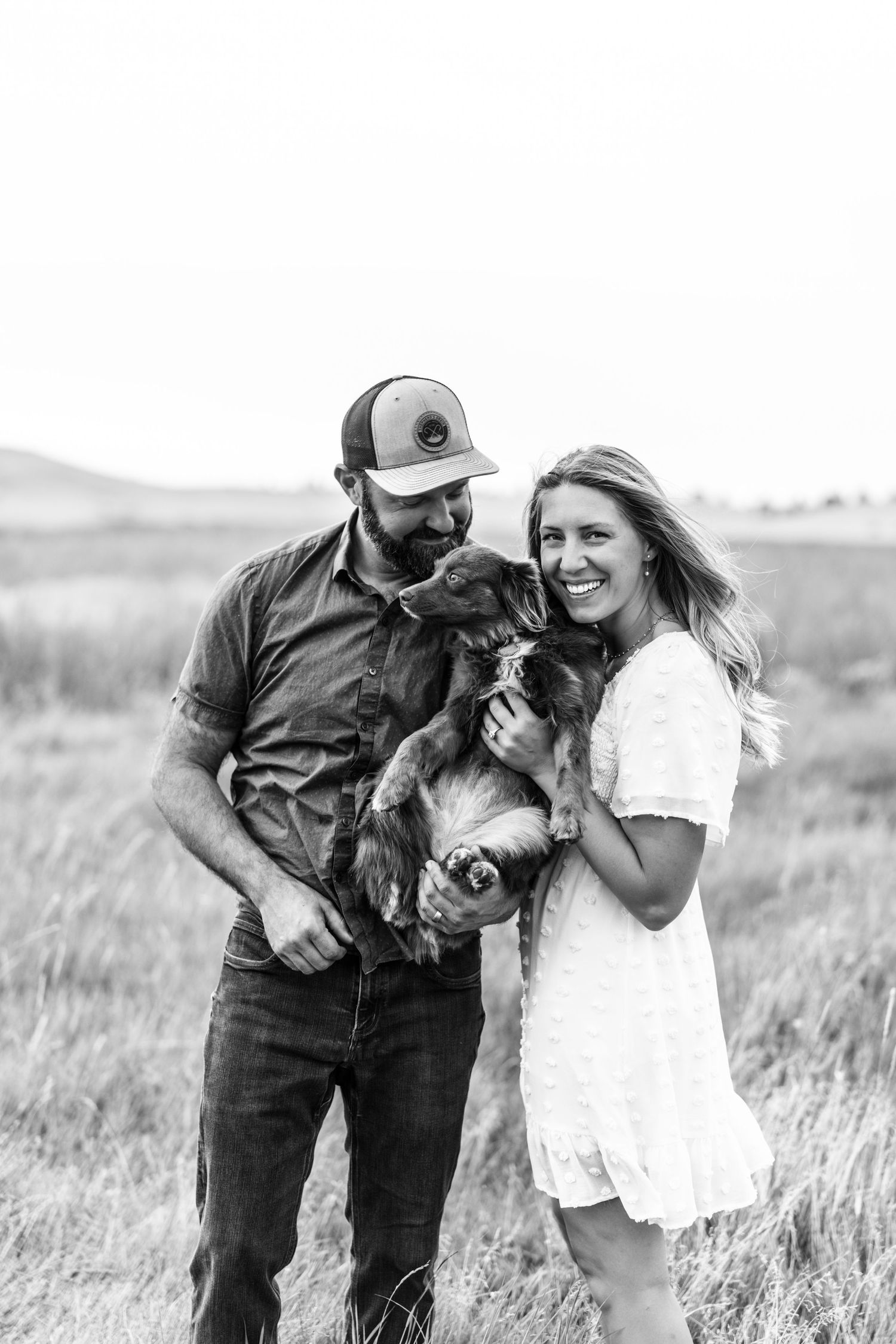 Smiling couple holding an adorable dog in a black and white outdoor field portrait.