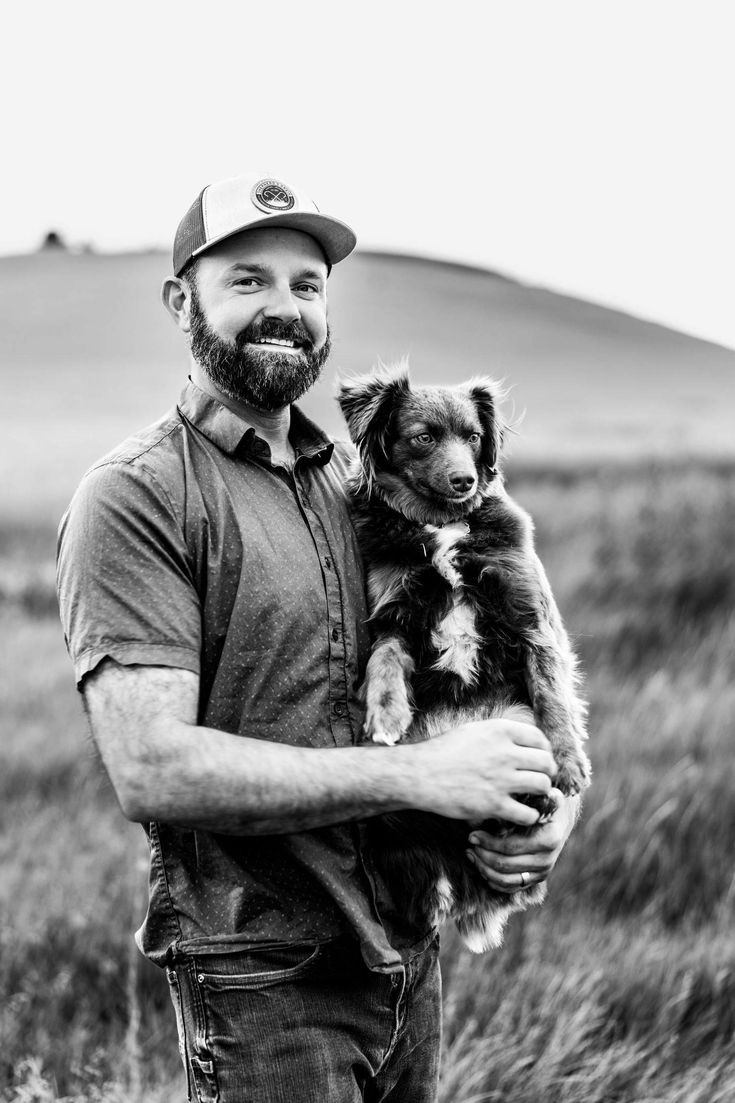 Bearded man in baseball cap holds fluffy puppy in black and white outdoor portrait with rolling hills.