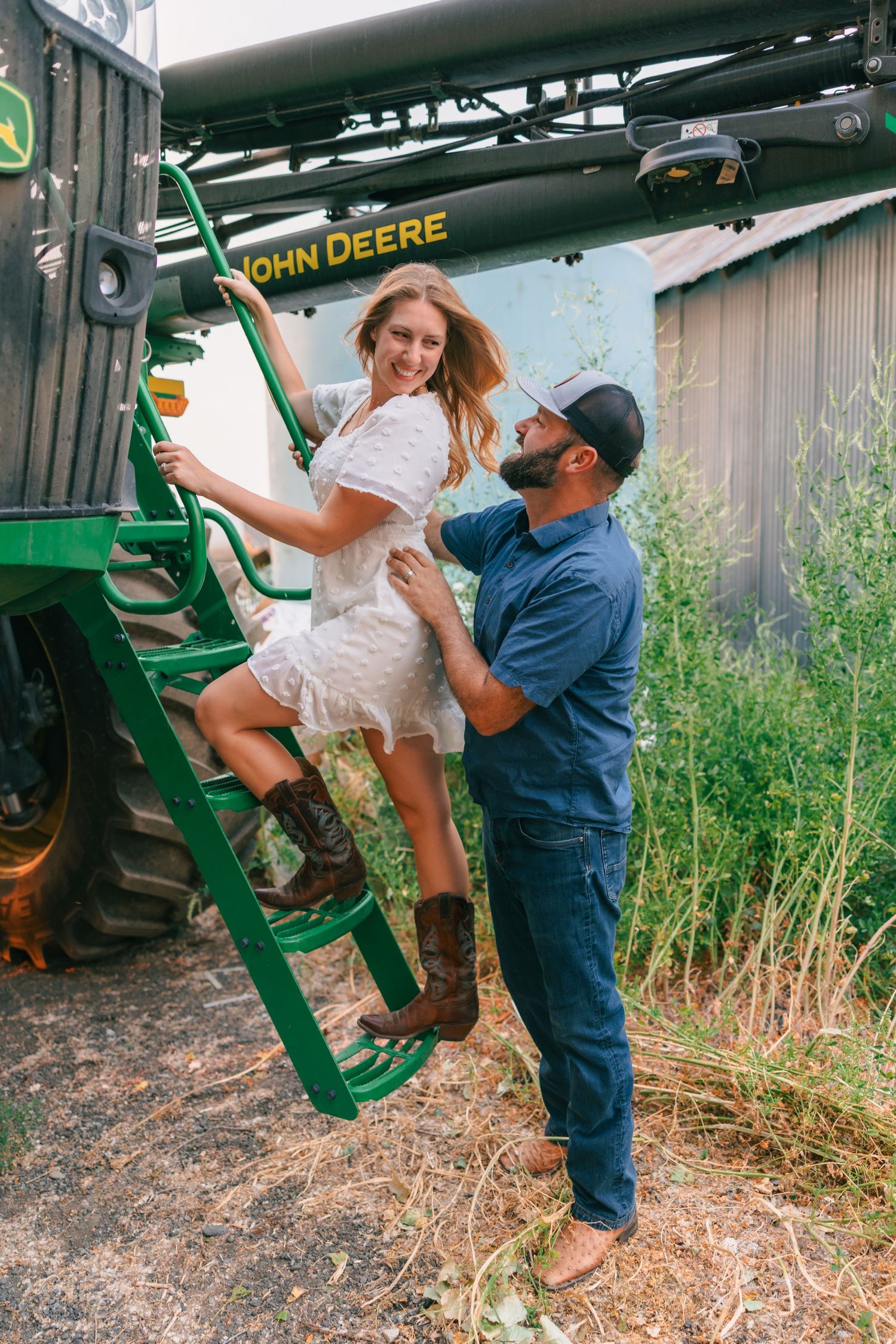 Couple posing with a John Deere combine harvester, woman in white dress on ladder, man in blue shirt assisting her.