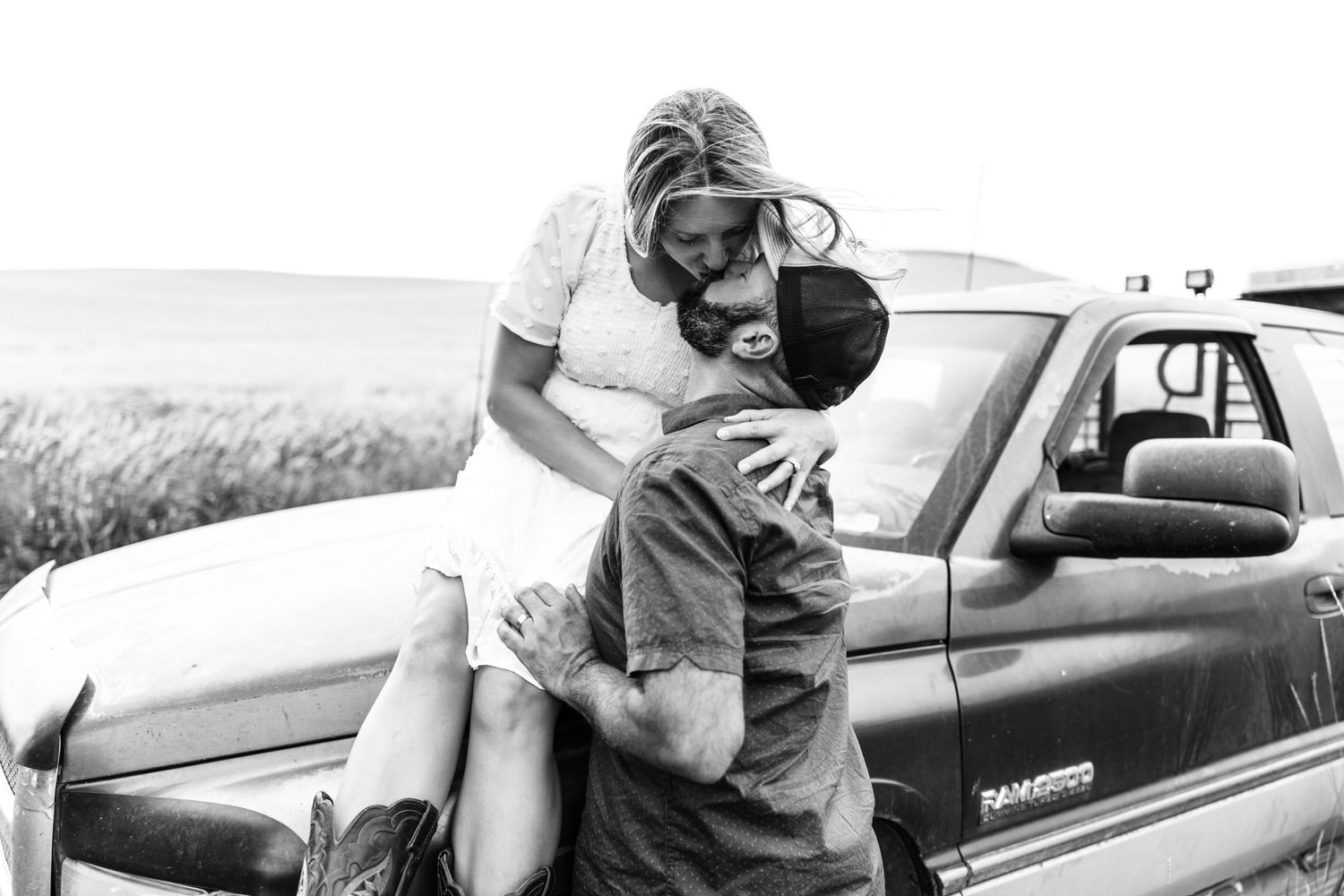 Black and white photo of a woman sitting on a truck bumper hugging a child in a rural field setting.