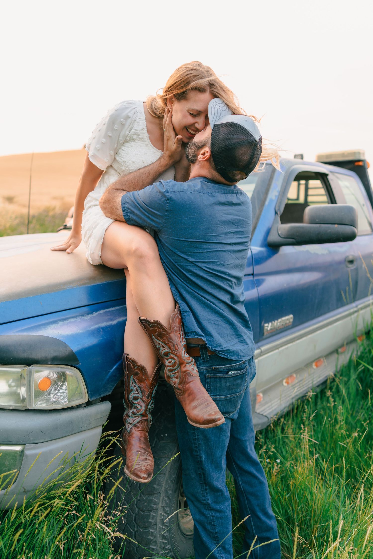 A woman in a white dress and cowboy boots sits on a blue truck hood as a man leans in for a kiss.
