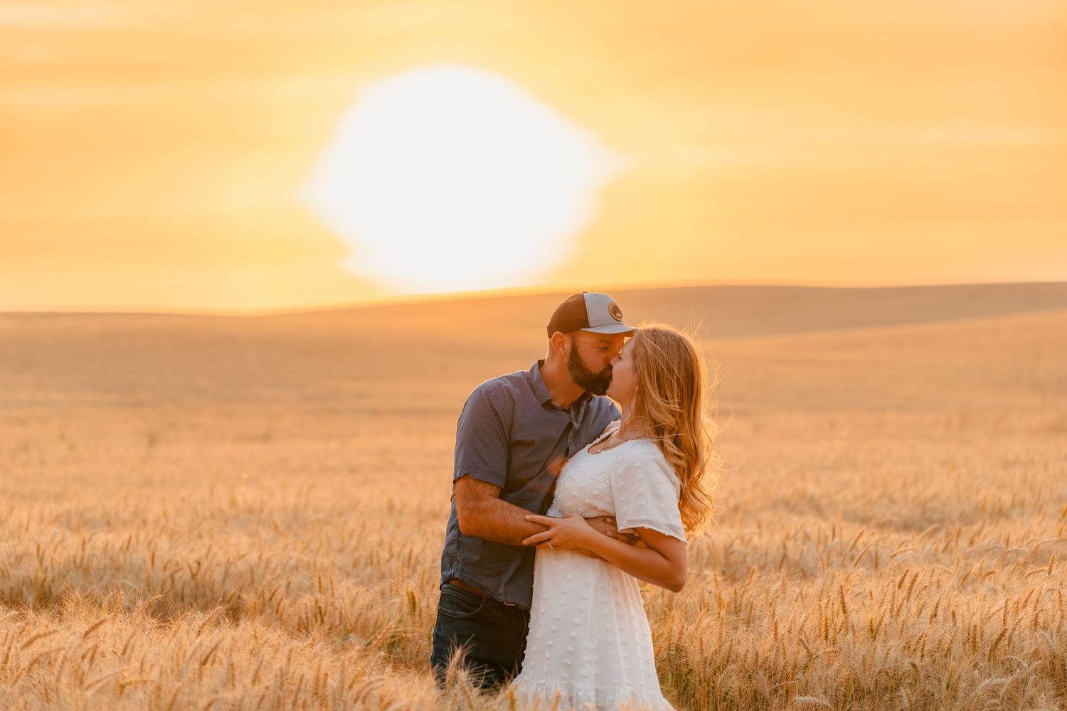 Couple sharing a romantic kiss at golden sunset in a vast wheat field, warm orange sky glowing behind them.