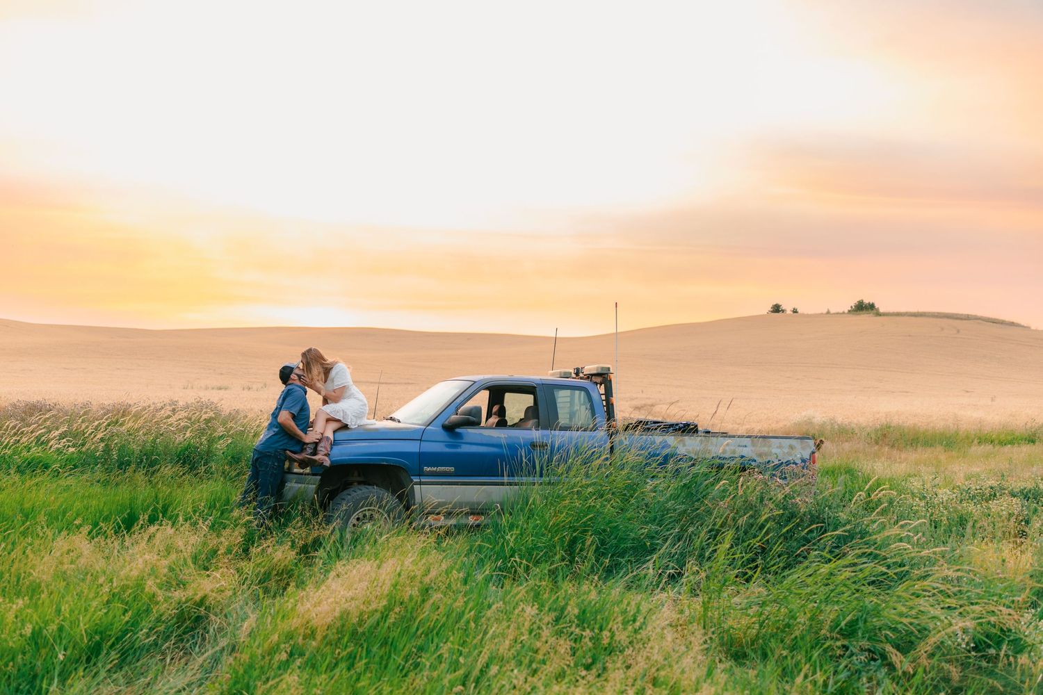 Couple sharing a romantic moment leaning against a vintage pickup truck in a golden sunset field.