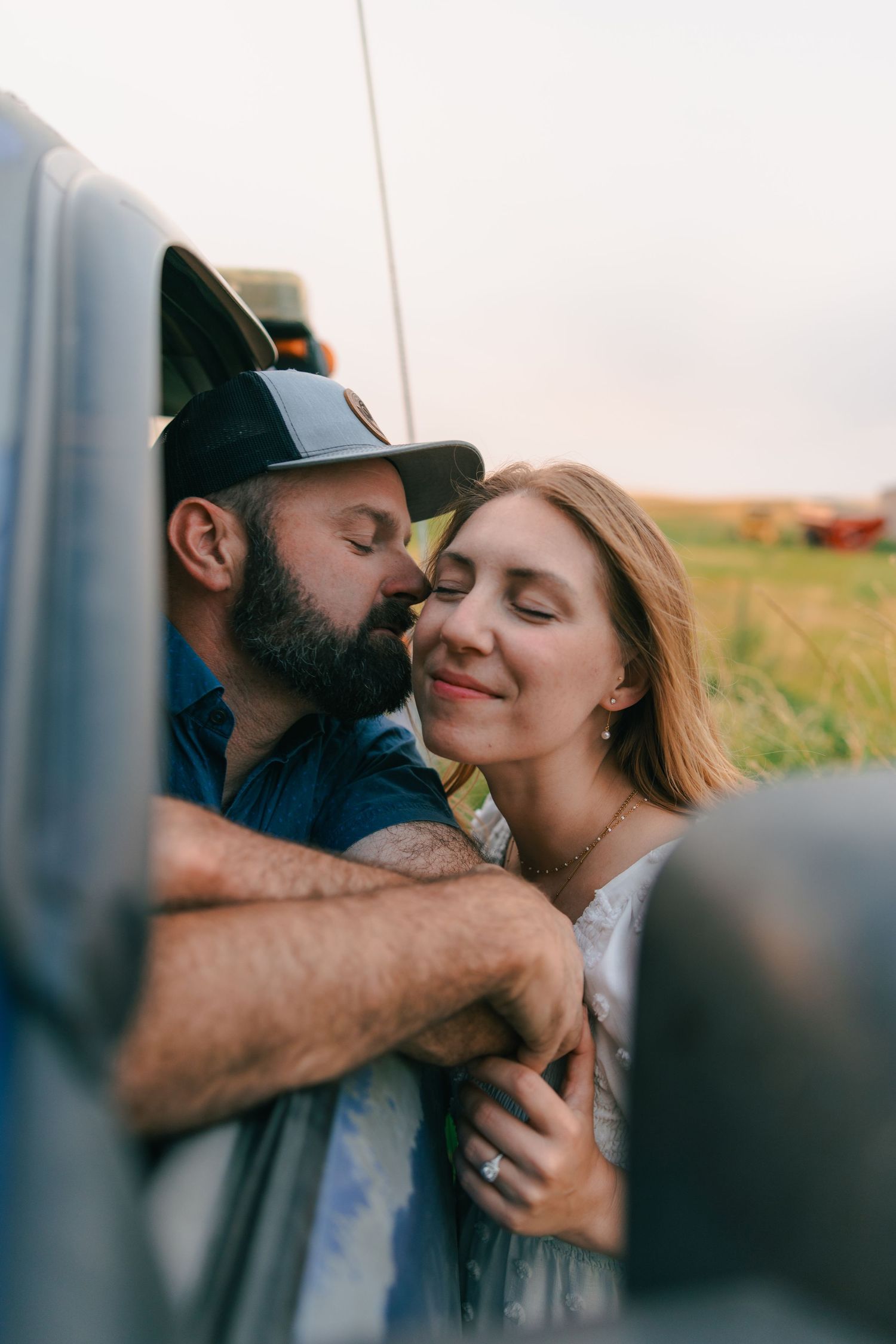 A bearded man in a cap kisses a smiling woman leaning against a truck window in a scenic field at sunset.