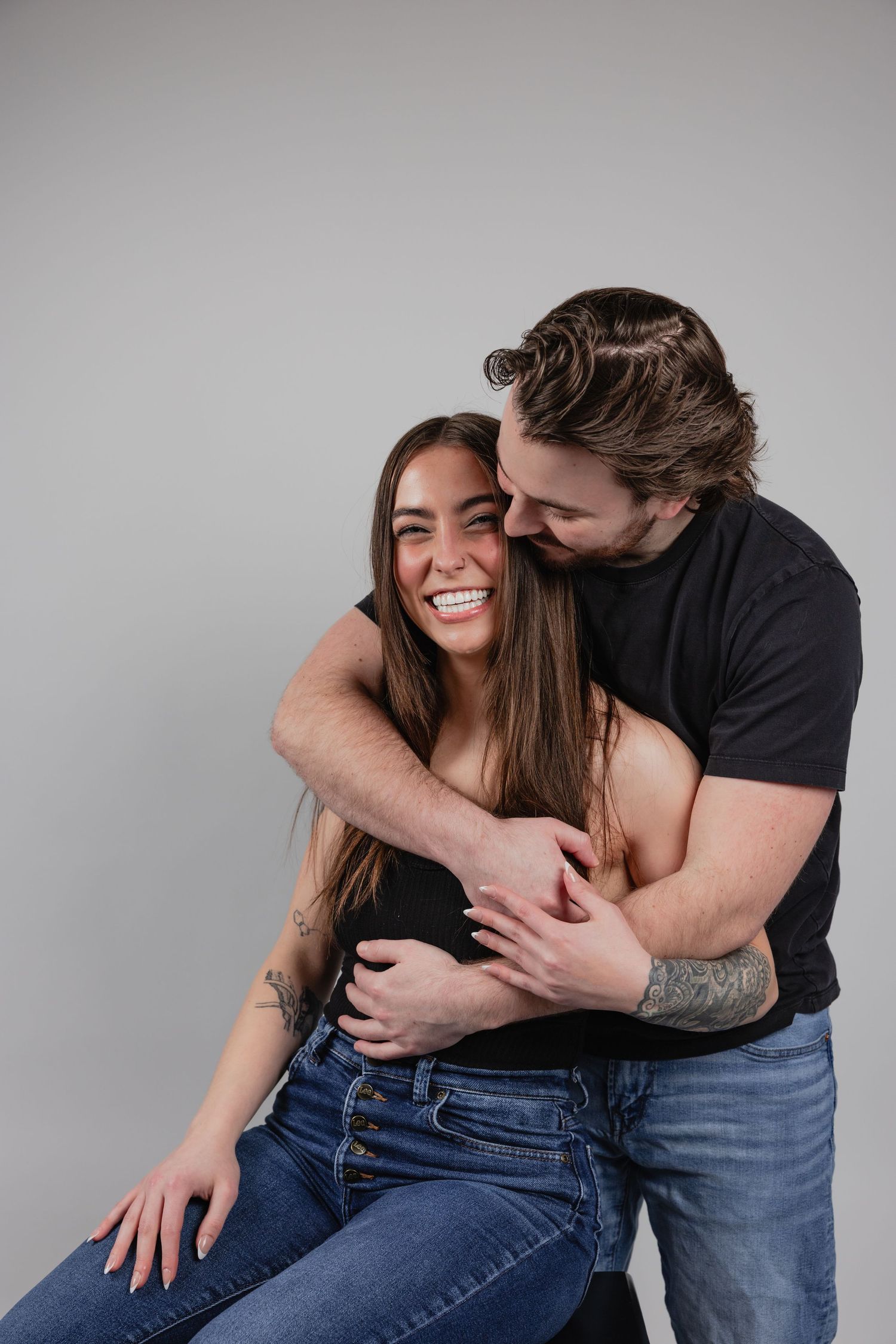 A playful couple embraces and laughs together during a casual photoshoot against a gray studio backdrop.
