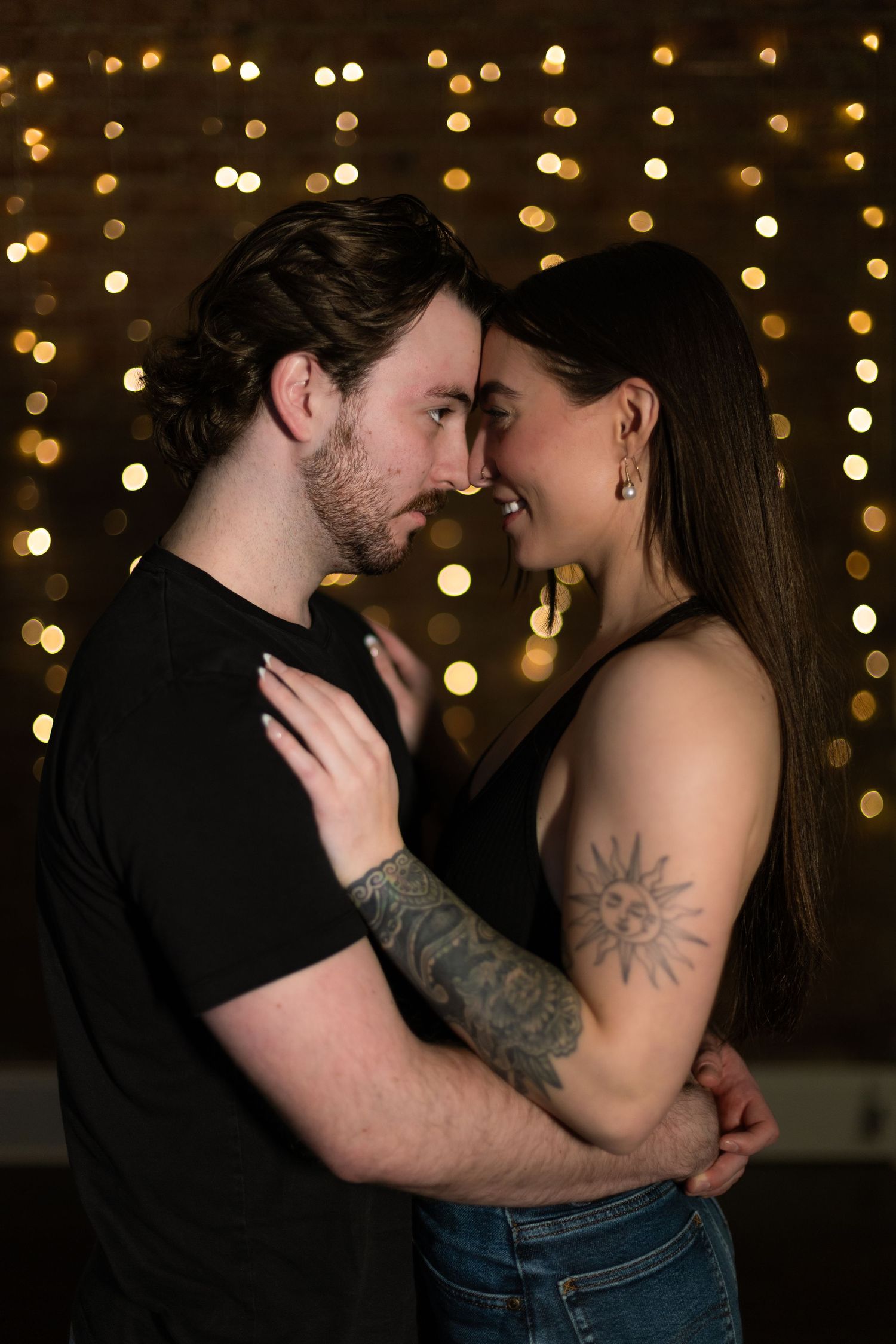 A romantic couple embraces in front of twinkling bokeh lights, with one showing a visible sun tattoo on their arm.