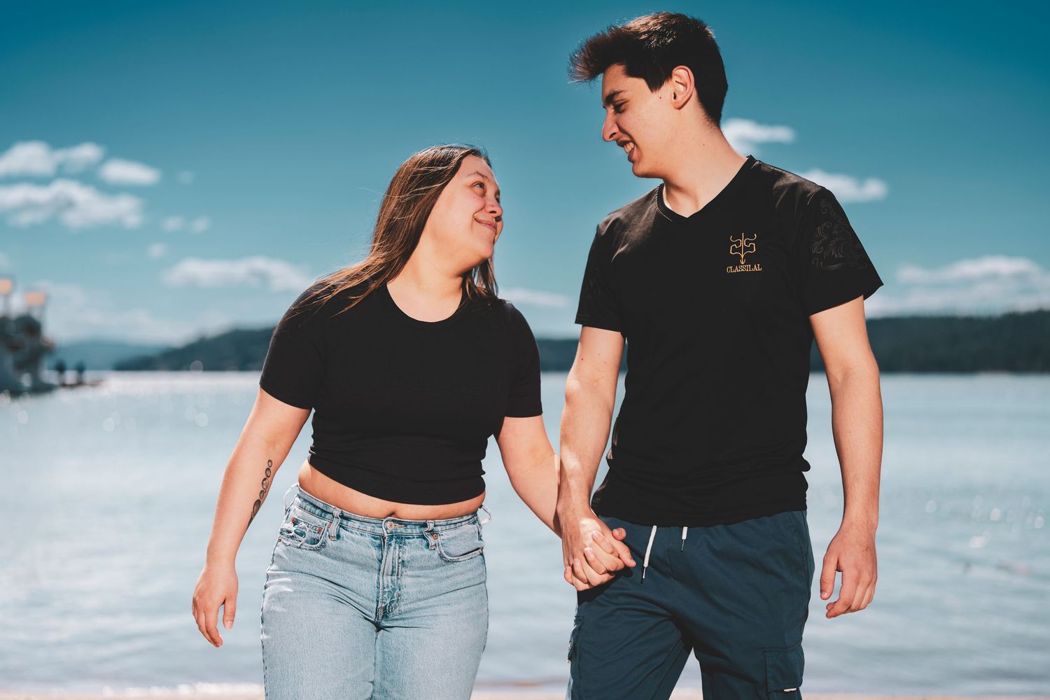 A couple in black shirts and jeans walk together on a beach with blue sky and water in the background.