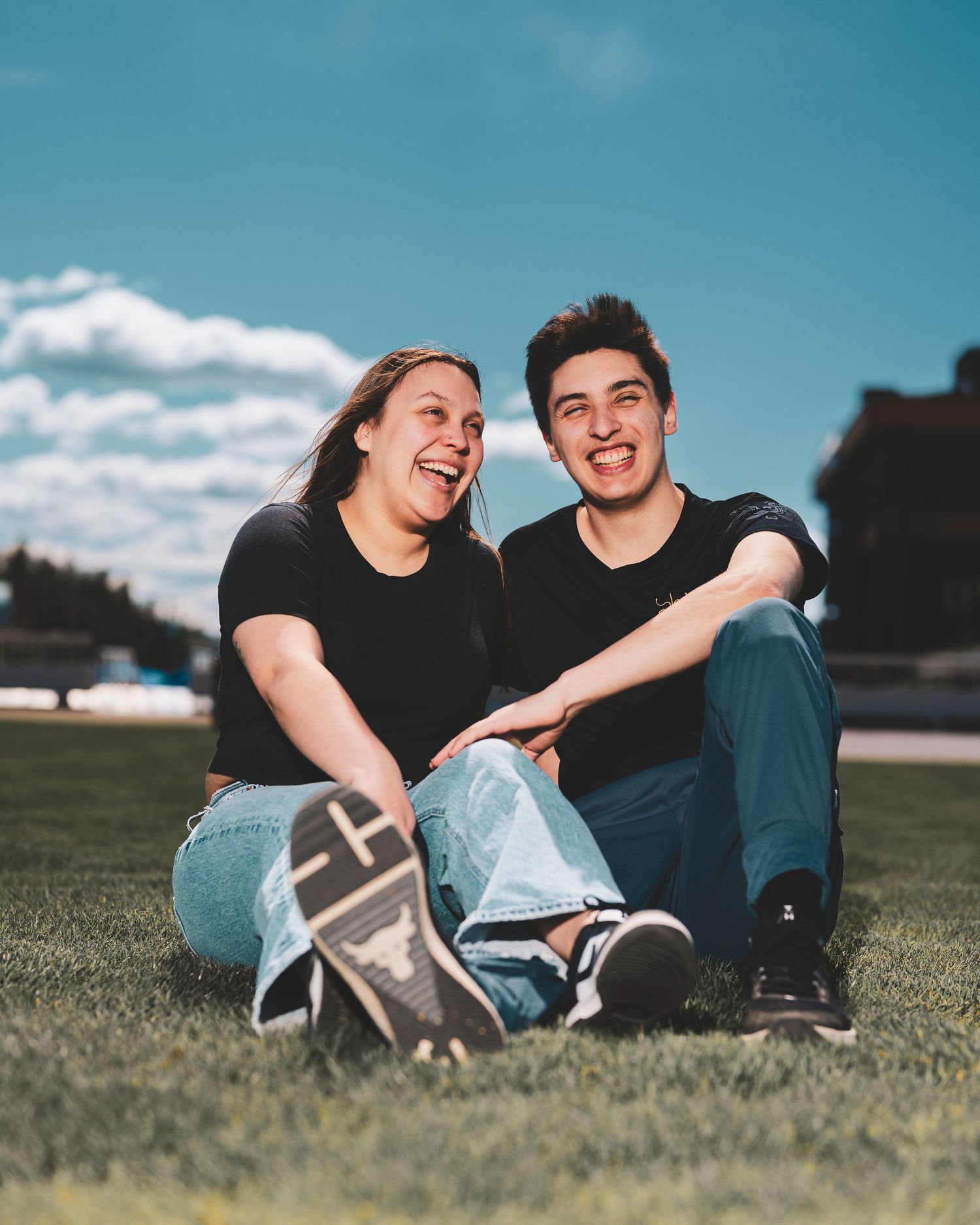 A couple in black shirts relaxes and laughs together while sitting on green grass on a sunny day.