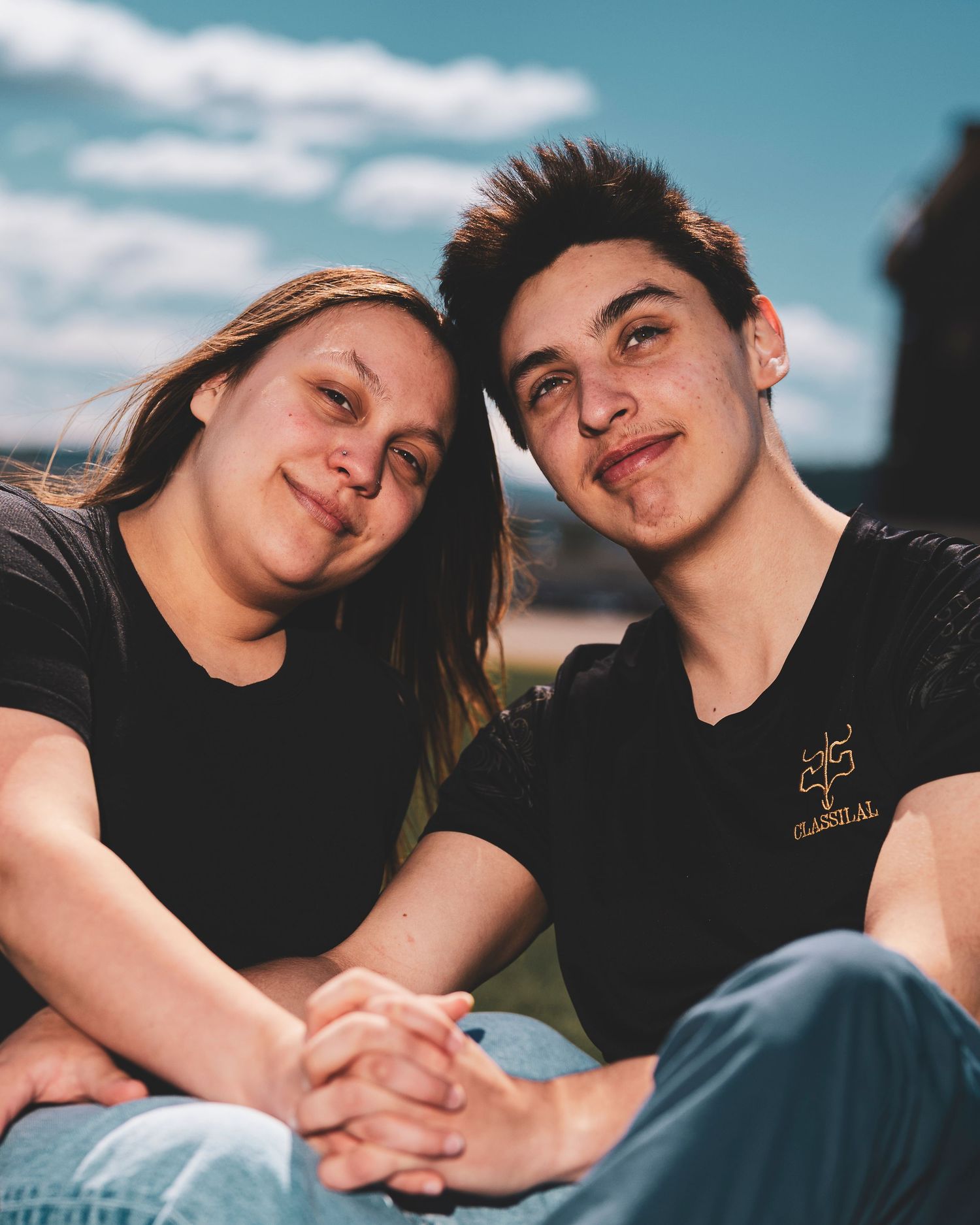 Two young people sitting close together holding hands and making playful expressions against a blue sky background.