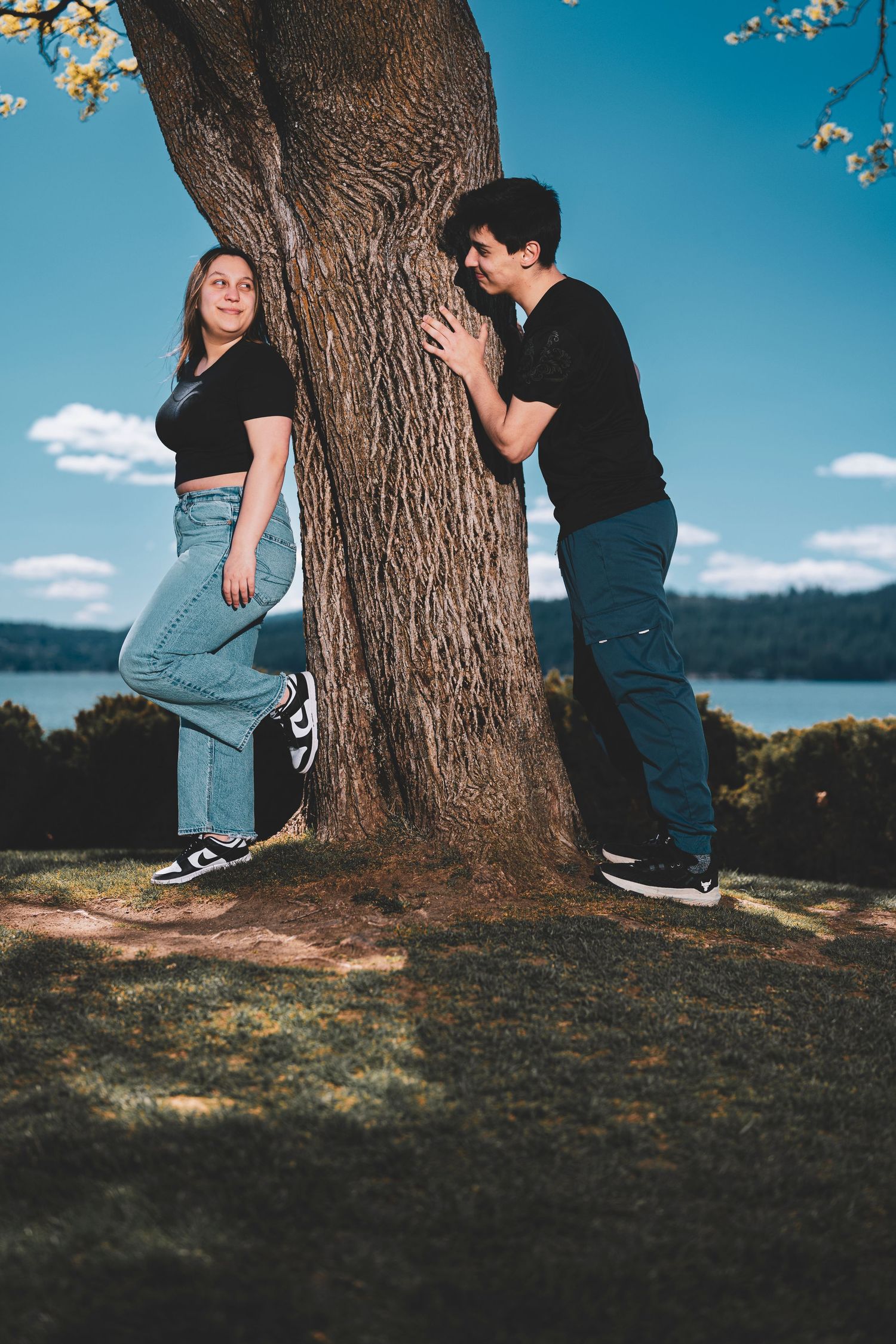 A couple stands behind a large tree trunk against a blue sky and lake backdrop in a romantic outdoor setting.