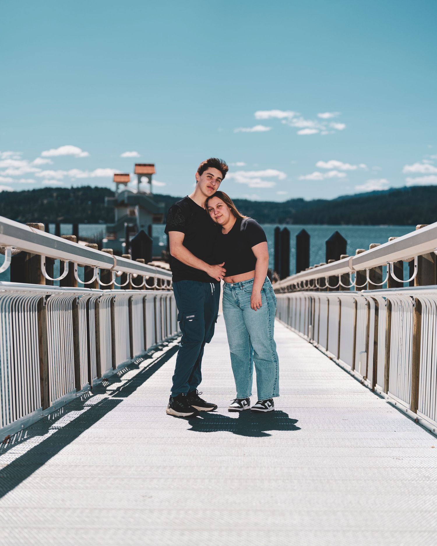 A couple embraces on a white bridge walkway with mountains and water visible in the background on a sunny day.