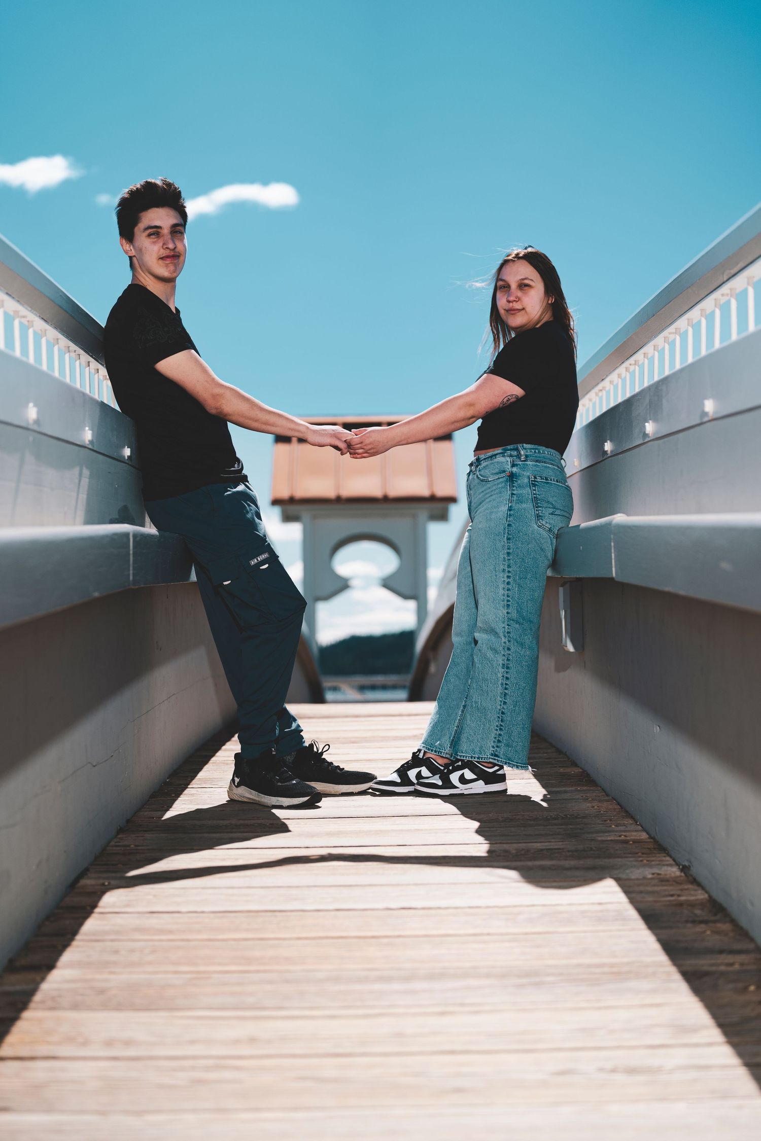 Two people in black shirts and jeans hold hands while standing on a wooden boardwalk under bright blue sky.