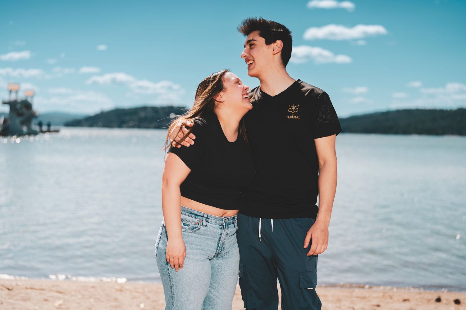Couple in casual outfits sharing playful moments on a scenic beach with blue water and sky in the background.