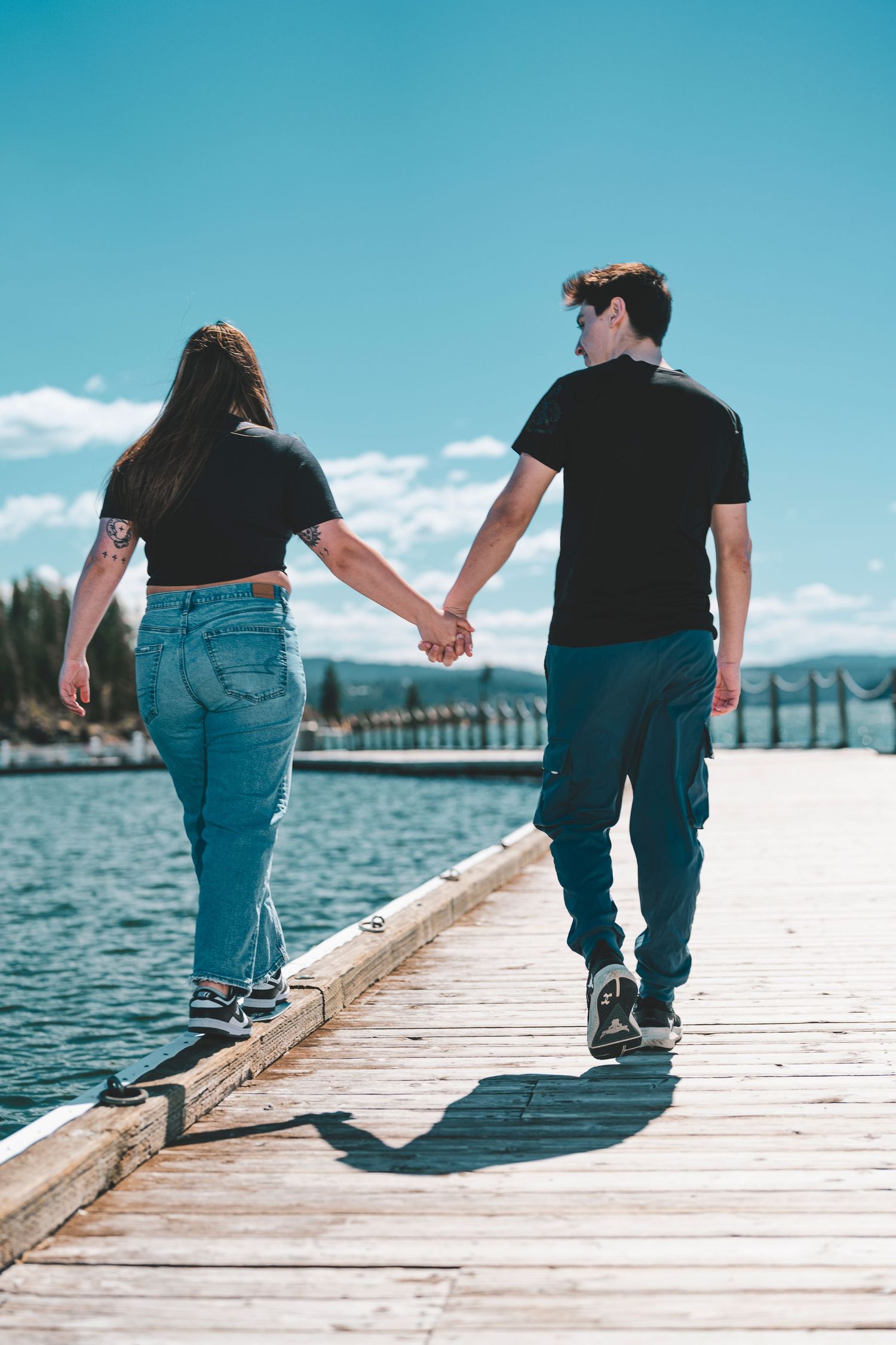 A couple holding hands while walking on a wooden pier by a lake on a sunny day with blue skies.