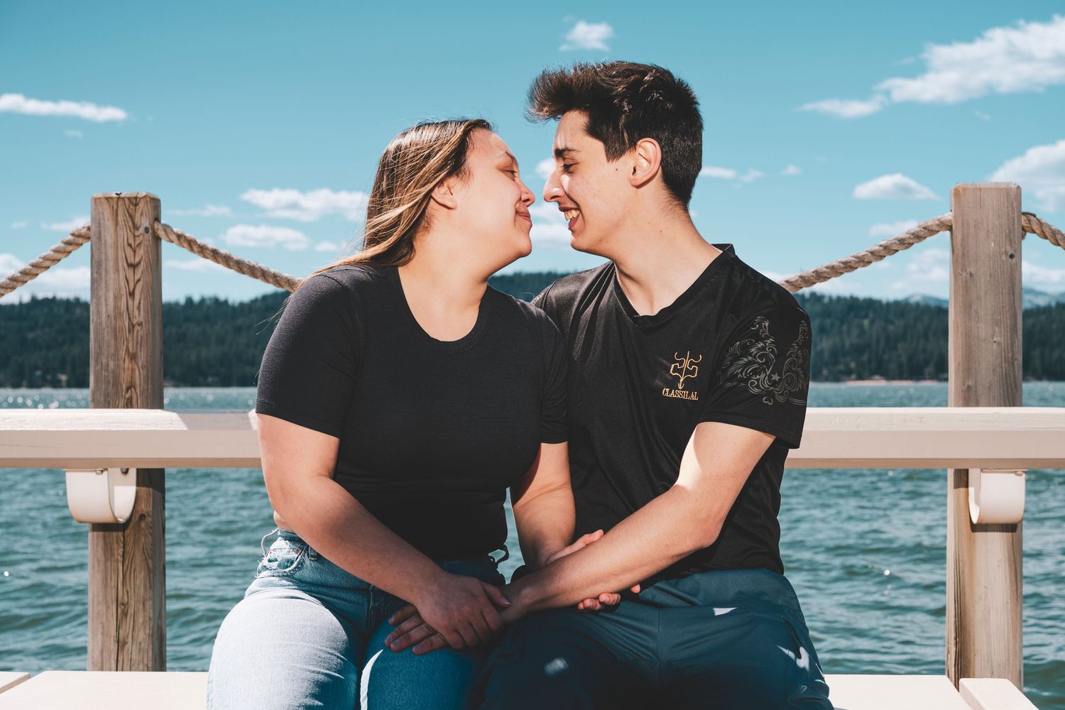 A couple in black shirts shares an intimate moment on a lakeside dock on a sunny summer day.