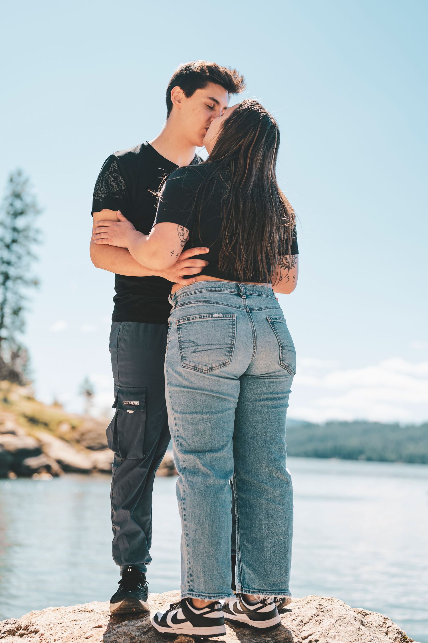 A couple embraces while standing by a scenic lakeside on a sunny day wearing casual denim and black clothing.