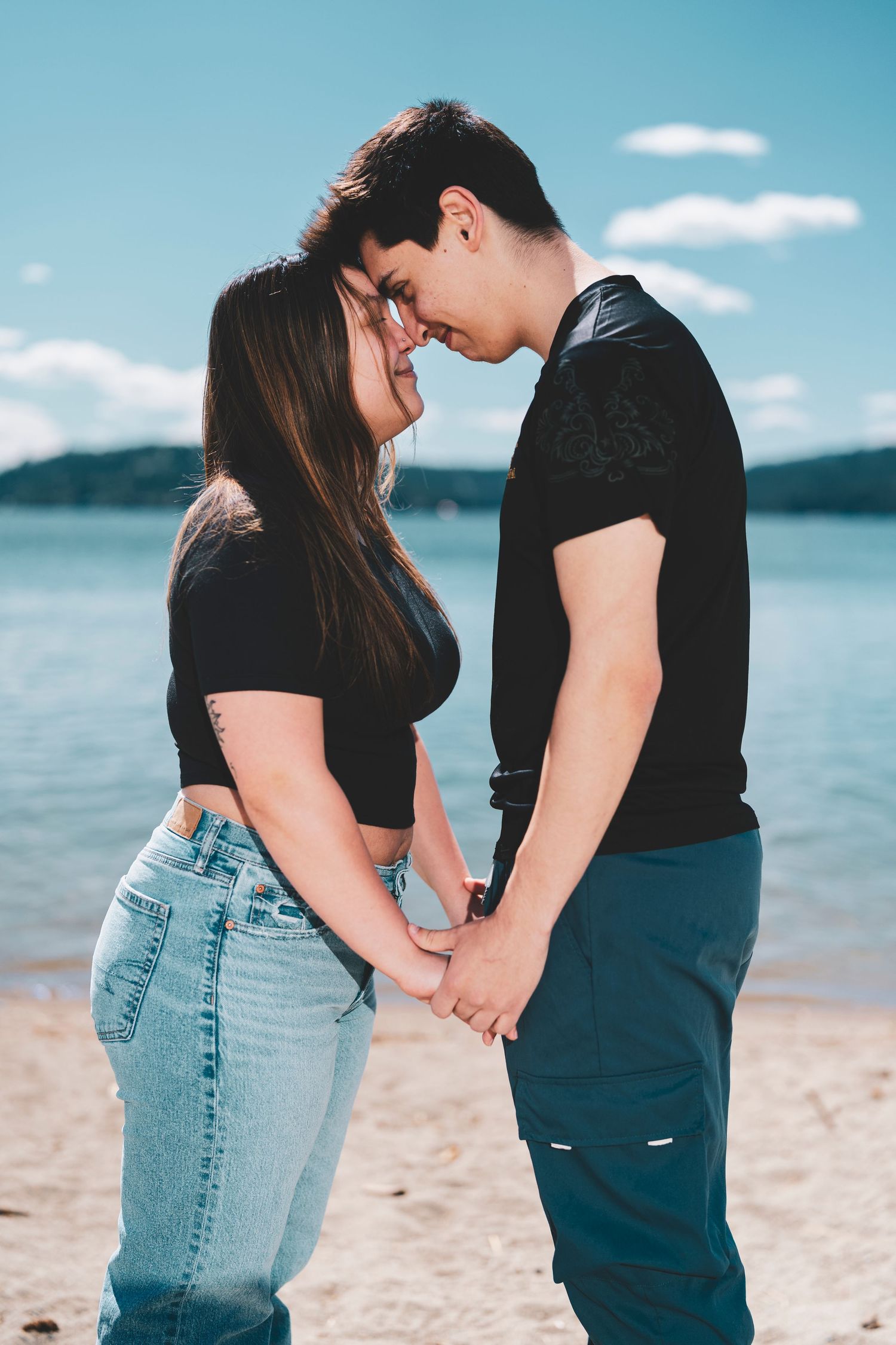 A couple wearing casual outfits stands close together on a sandy beach with blue water and sky in the background.
