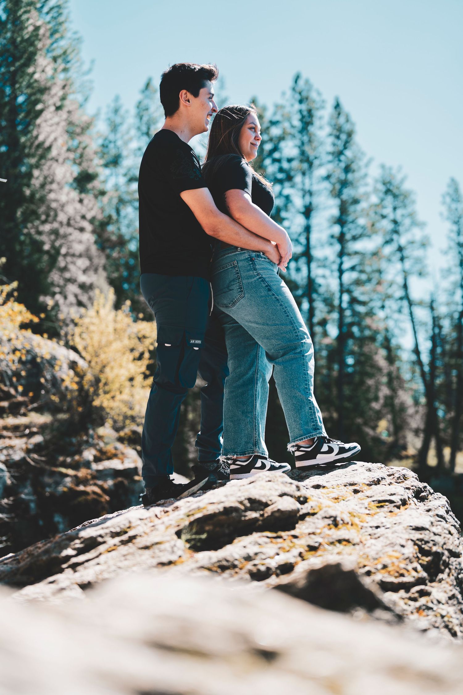 Two people sit close together on a rocky overlook surrounded by evergreen trees in a peaceful mountain setting.