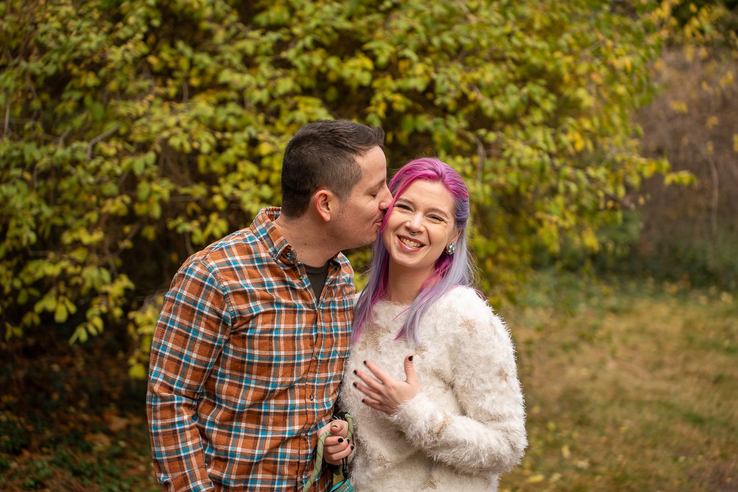 A man in a plaid shirt kisses a smiling woman with purple hair wearing a fuzzy white sweater outdoors in autumn.