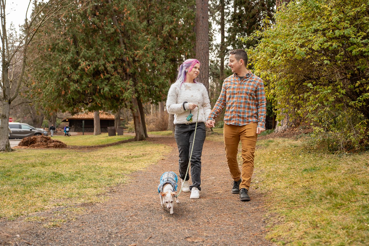 Couple holding hands walking their small dog on a gravel path in a lush green park.