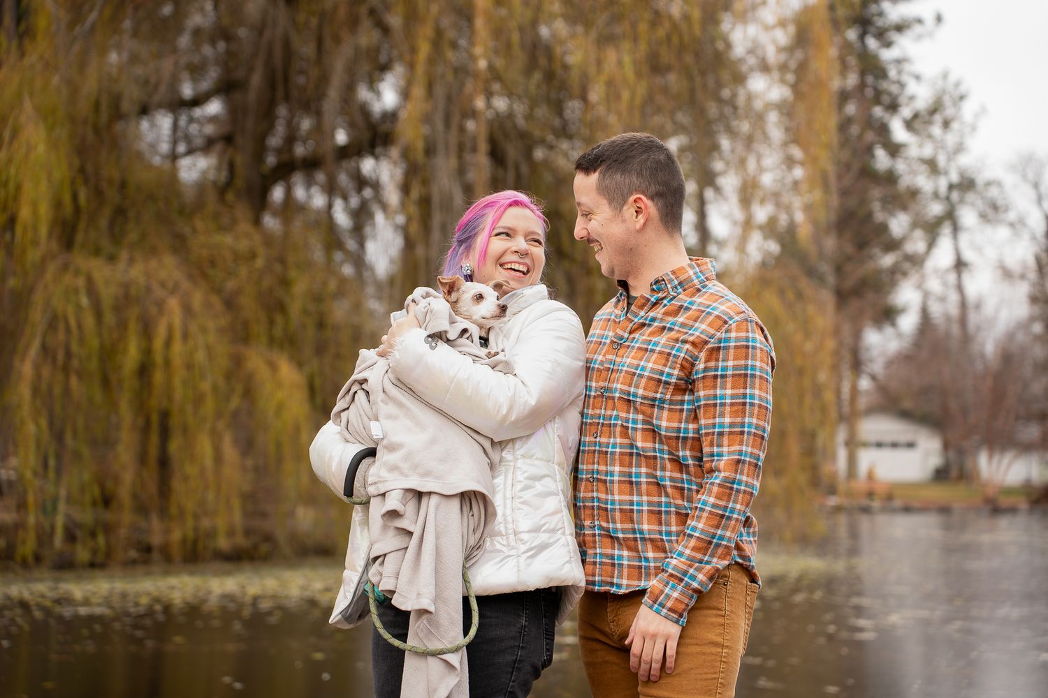 Couple smiling outdoors by a lake in autumn, woman with pink hair holding a small dog wrapped in a blanket.