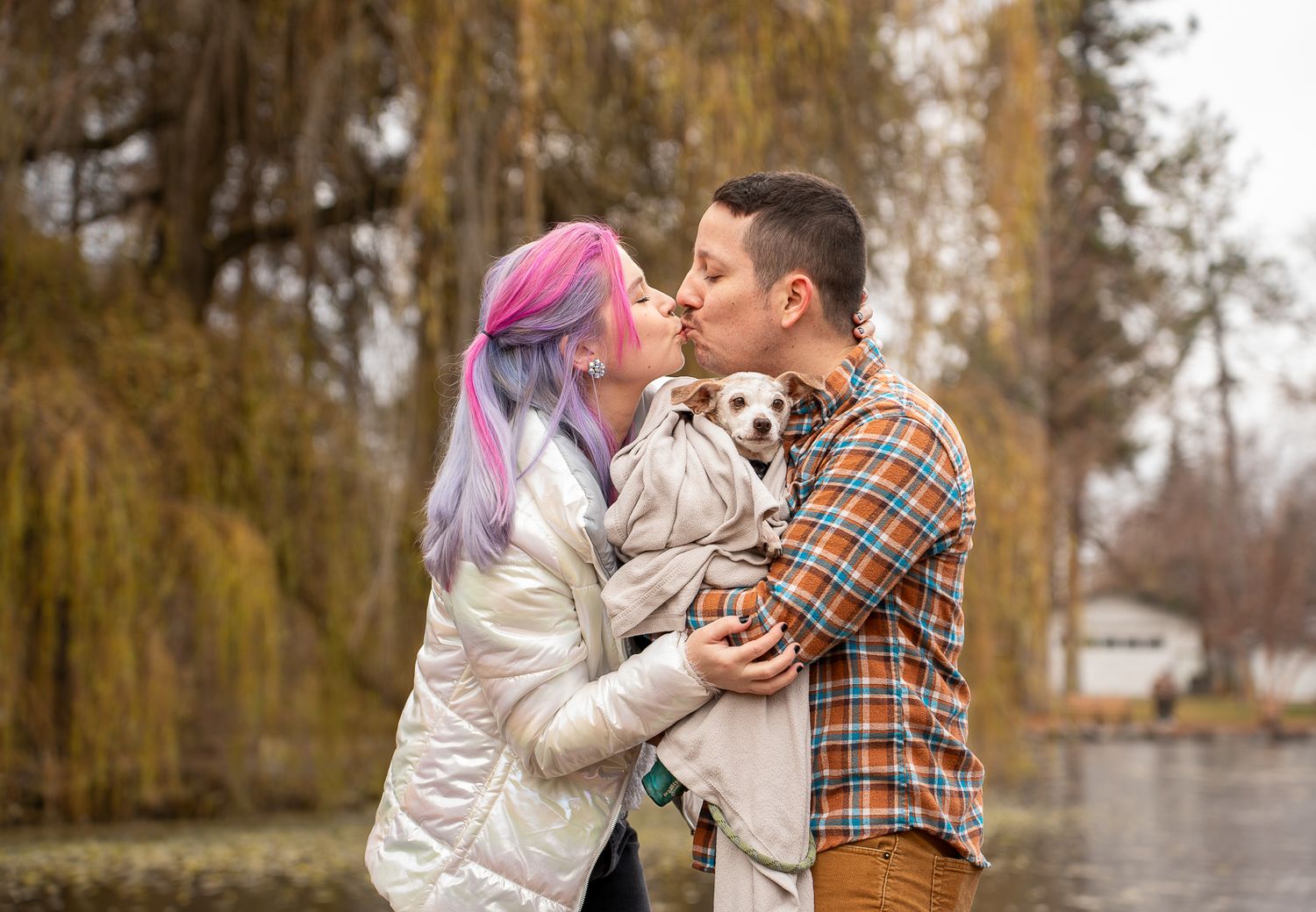 Couple kissing while holding a small dog outdoors near a lake with willow trees in autumn.
