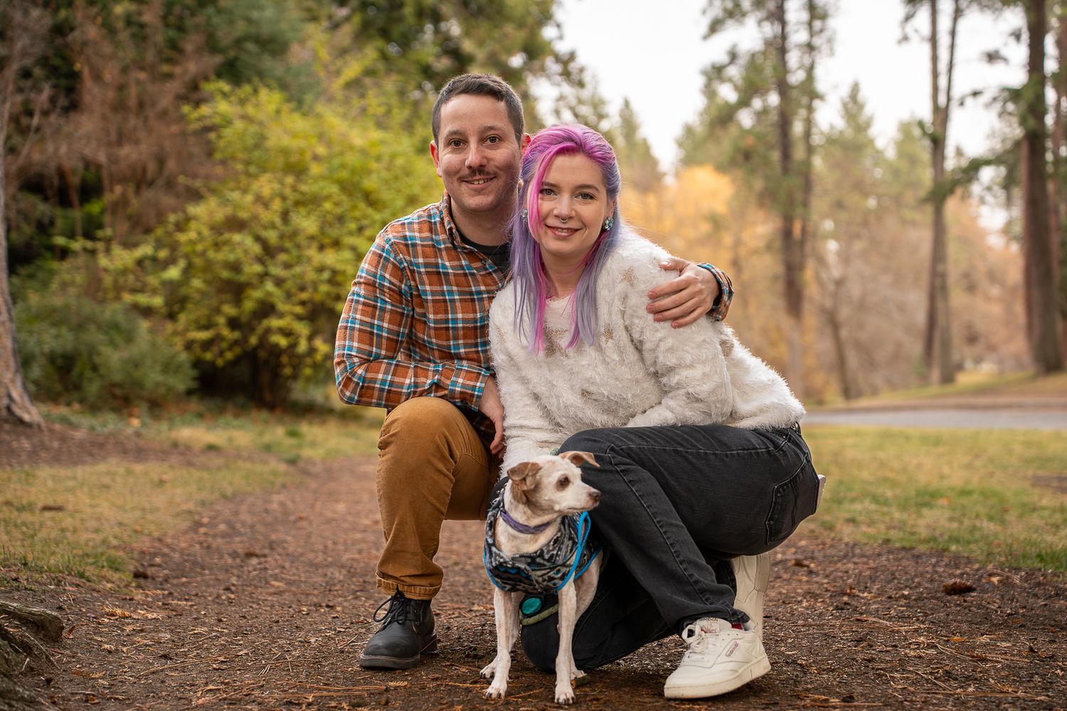 Couple with purple-haired woman and man in plaid shirt pose with small dog on forest trail in autumn.