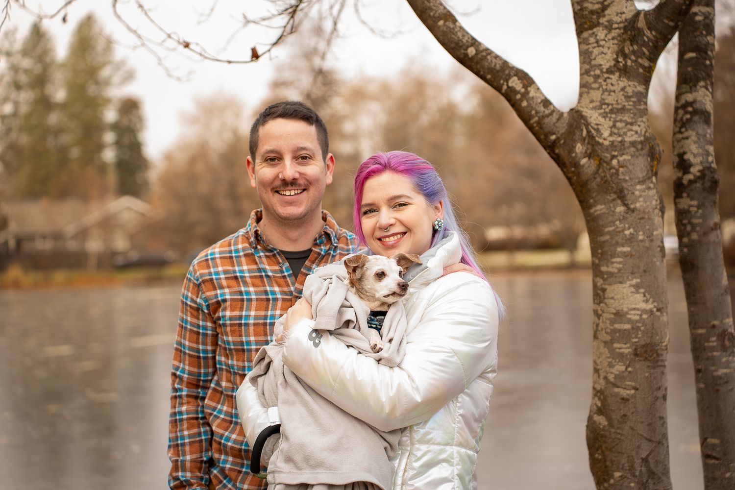 Smiling couple posing outdoors by a lake, woman with pink hair holding a small dog.