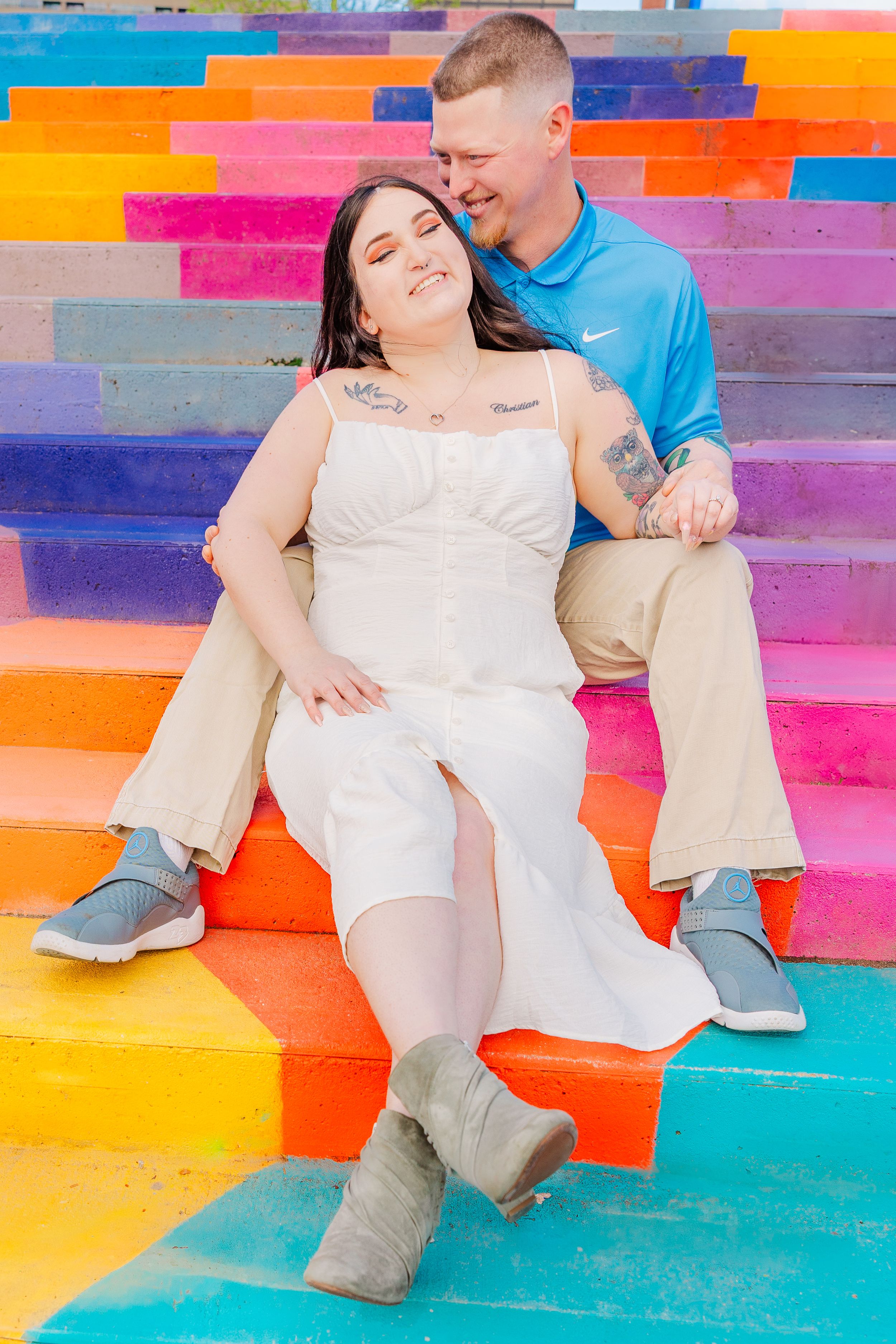 Couple sits together on vibrant rainbow-colored stairs, wearing white dress and blue shirt.