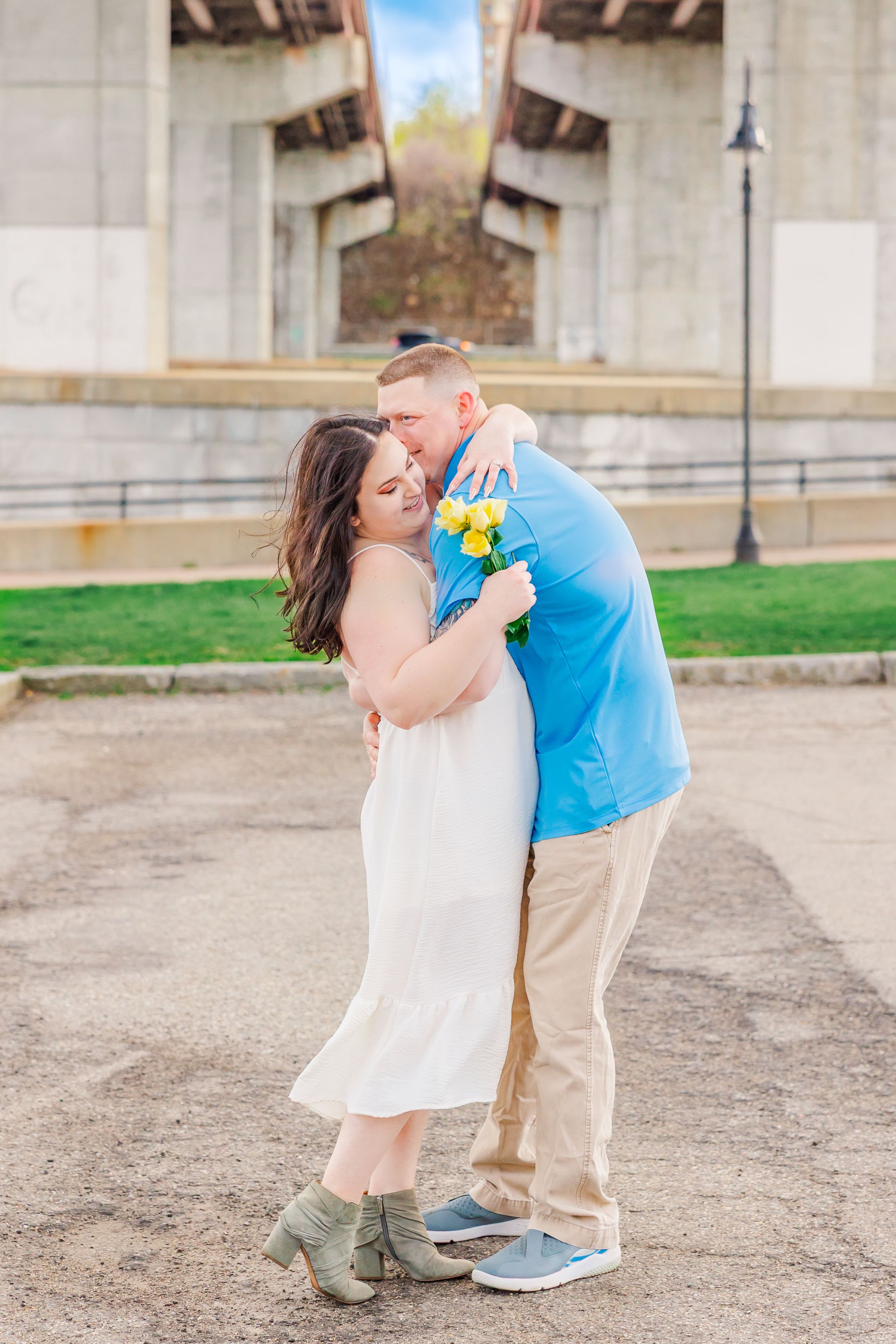 A couple shares a romantic moment under a concrete bridge while holding yellow flowers.