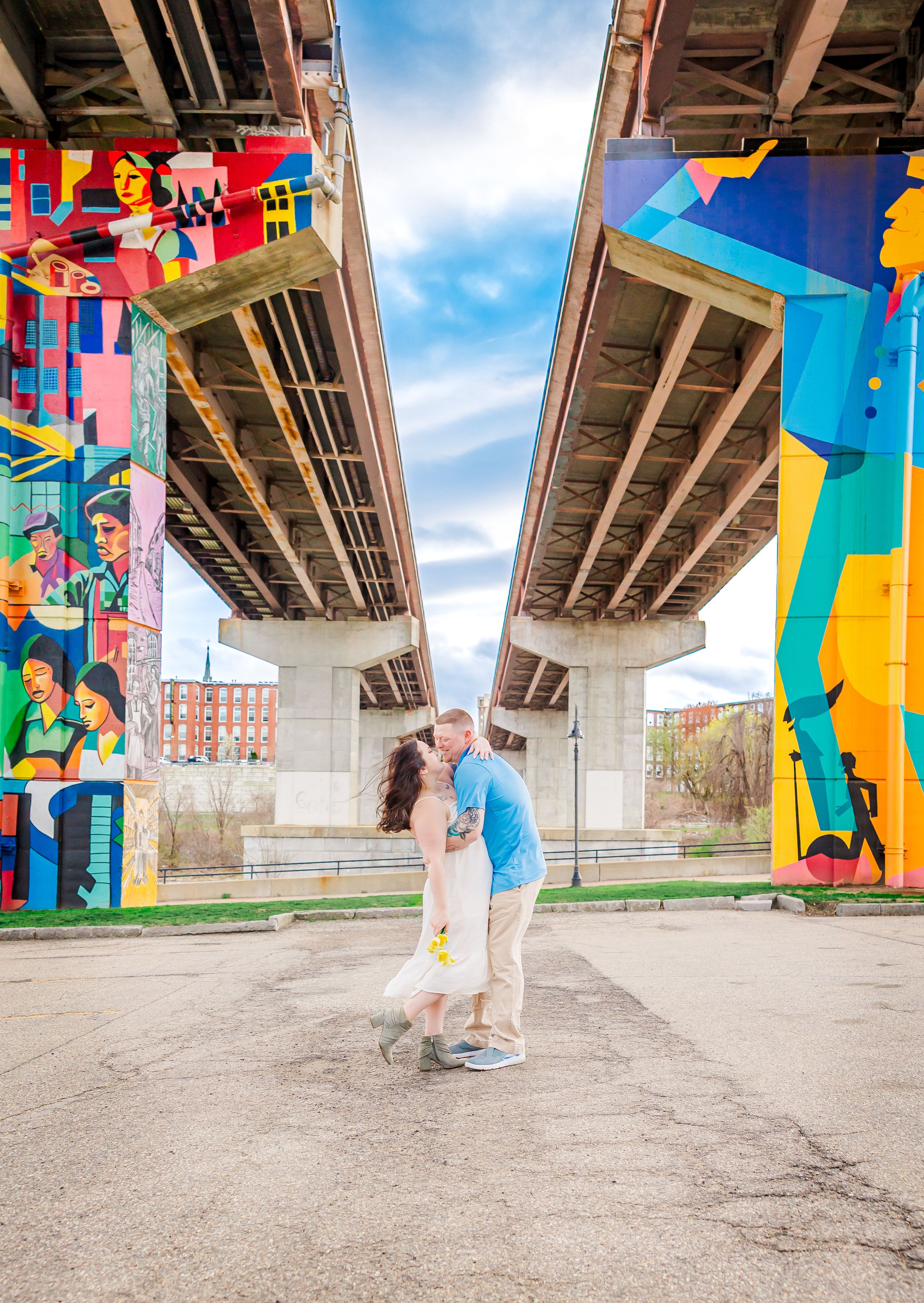 A couple embraces under a bridge with vibrant colorful murals painted on the concrete supports.