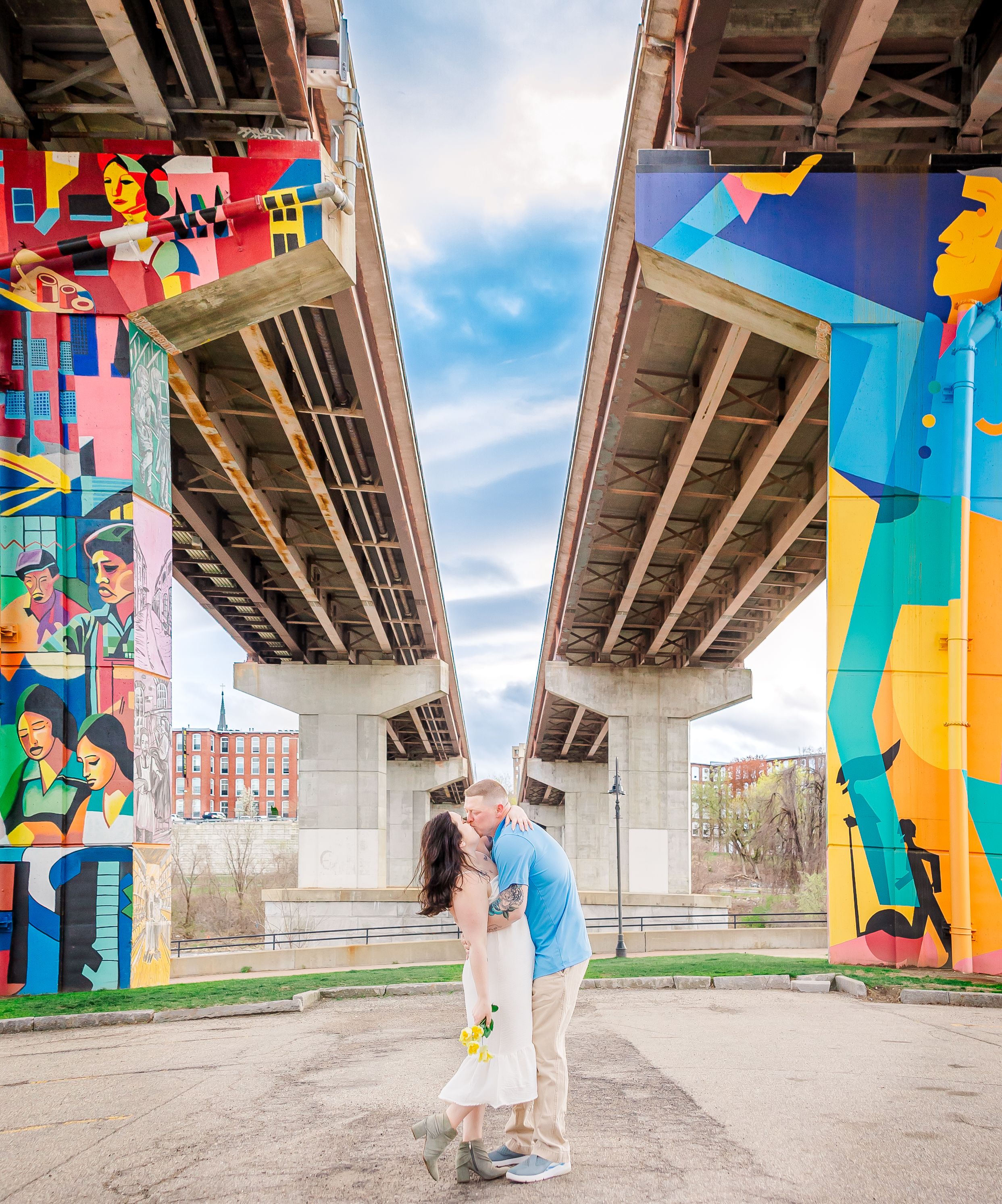 Couple kissing under bridge with colorful street art murals and blue sky.