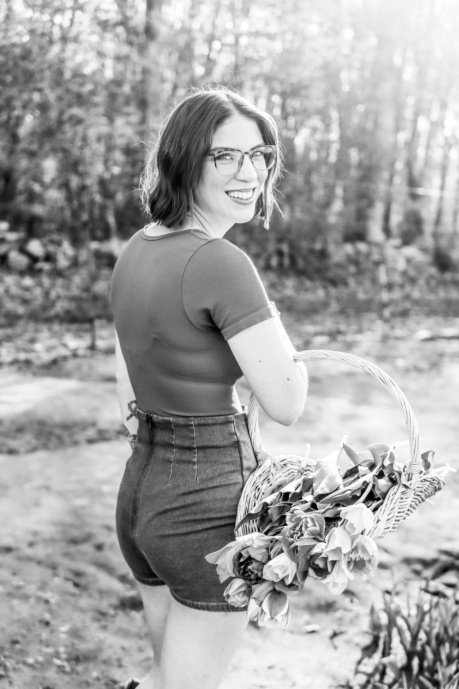 Monochrome image of a girl standing in a flower field and holding a basket of tulips while facing the camera.