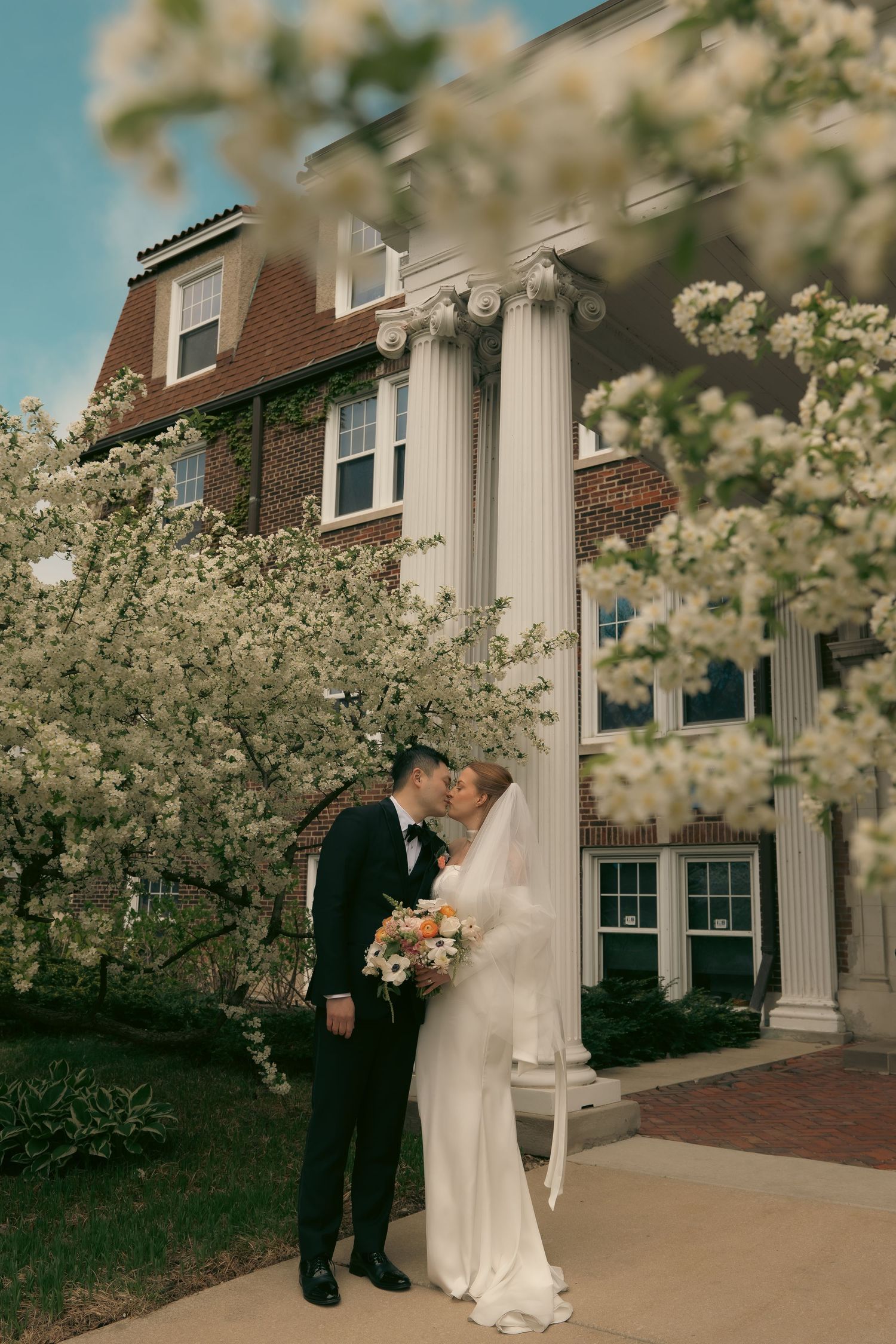 A romantic wedding moment captured in spring with blooming cherry blossoms and classic brick colonial architecture.