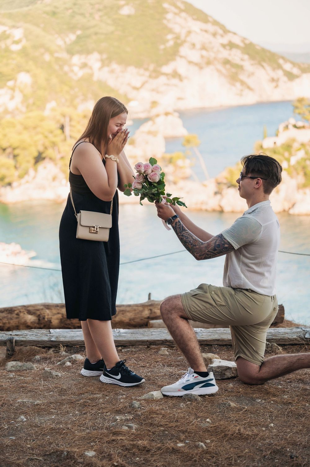 Romantic proposal scene with person kneeling on coastal viewpoint holding flowers.
