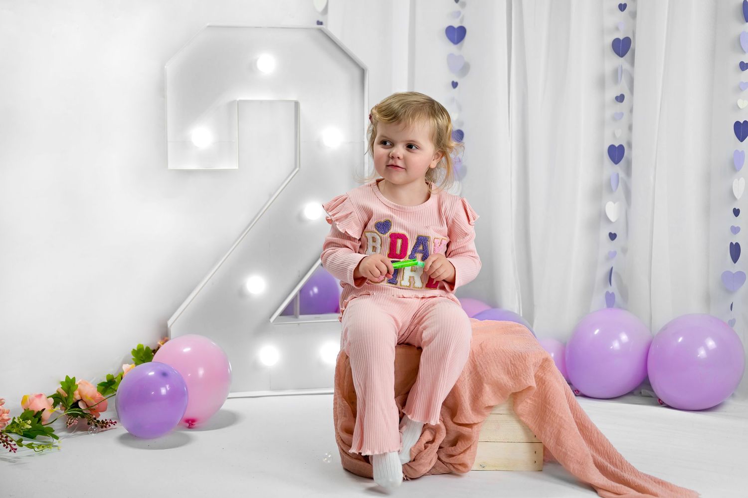 A child sits in a pink chair at a 2nd birthday party setup with purple and pink balloons and floral decorations.