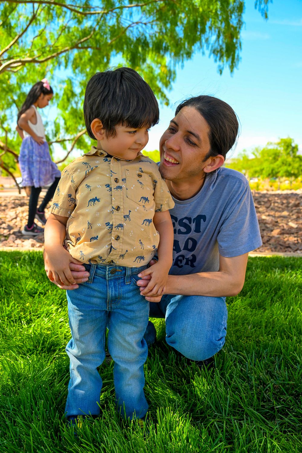 A father kneels on green grass smiling at his young son in a dinosaur shirt and jeans at an outdoor park.