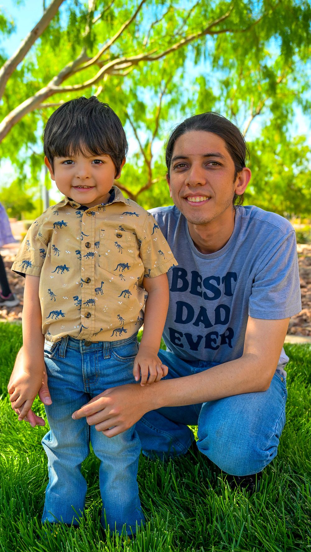 A young boy in a dinosaur shirt poses with his smiling father wearing a Best Dad Ever t-shirt outdoors.