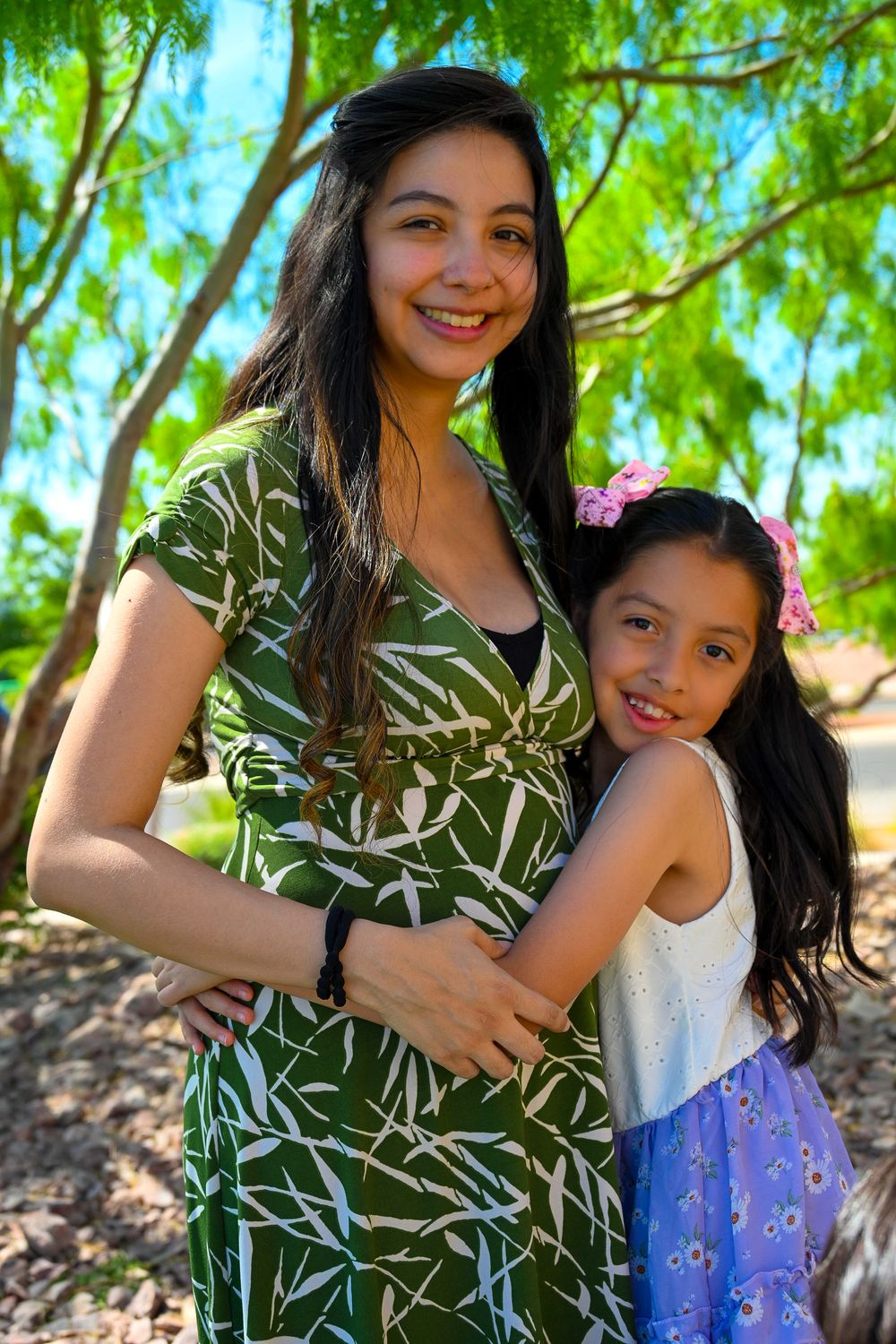 A smiling pregnant woman in a green dress hugs her young daughter with a pink flower in her hair outdoors.