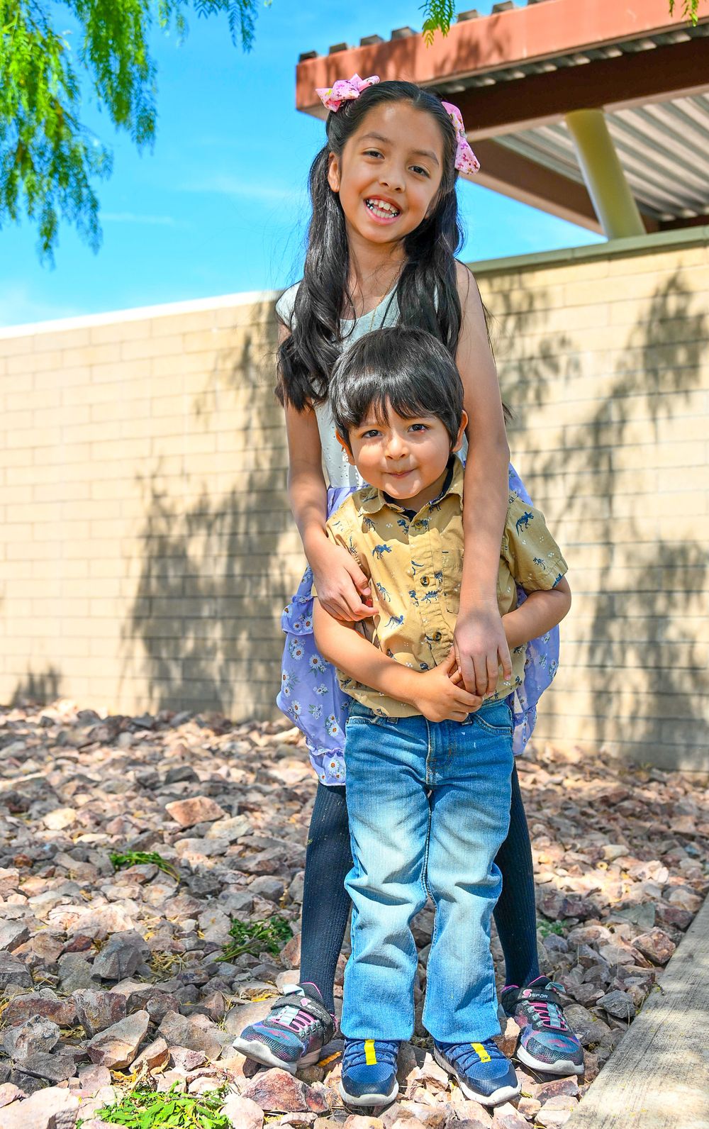 A young girl hugs her little brother from behind as they pose together smiling outdoors near a brick wall.
