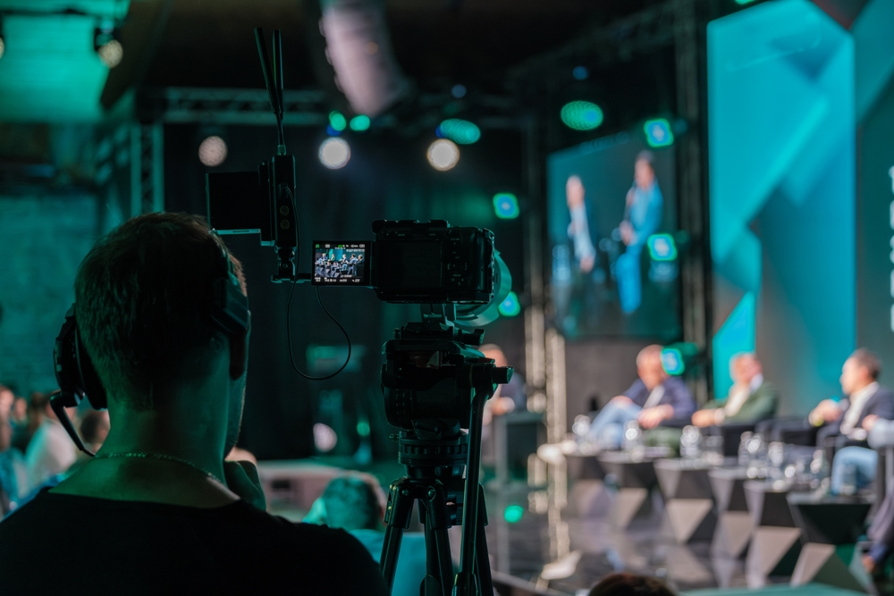 A camera operator films a panel discussion on stage with turquoise lighting in the background.