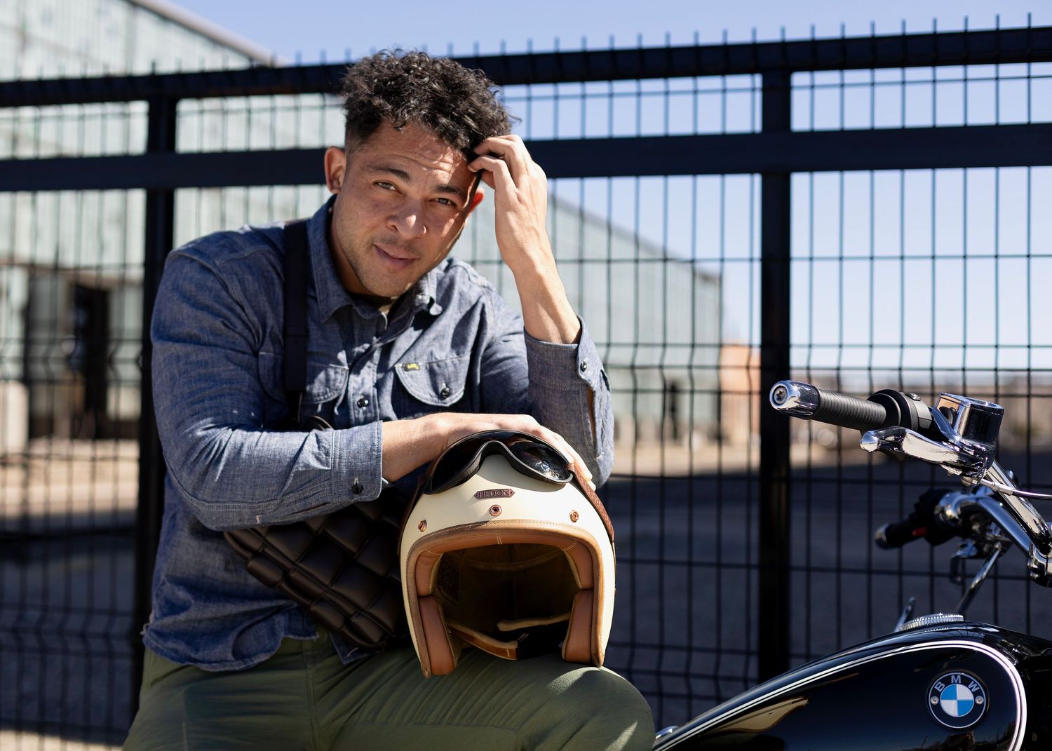 A motorcyclist rests against a fence while holding a vintage tan motorcycle helmet.