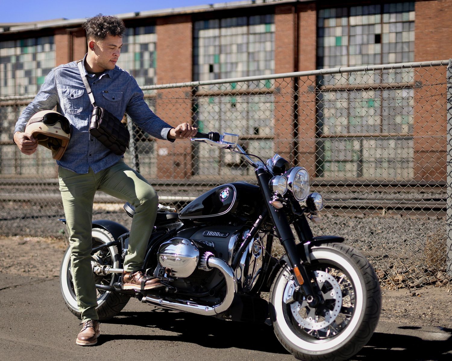 Motorcyclist poses with classic black BMW motorcycle near industrial brick building.