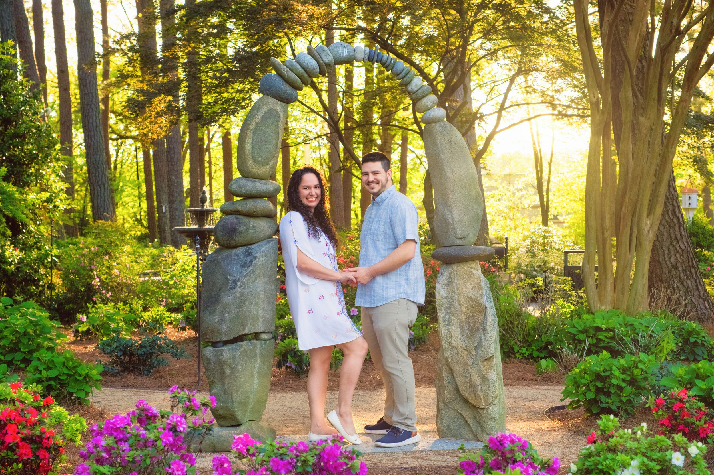 A couple stands under a rustic stone arch surrounded by vibrant pink flowers and sunlit trees in a garden setting.