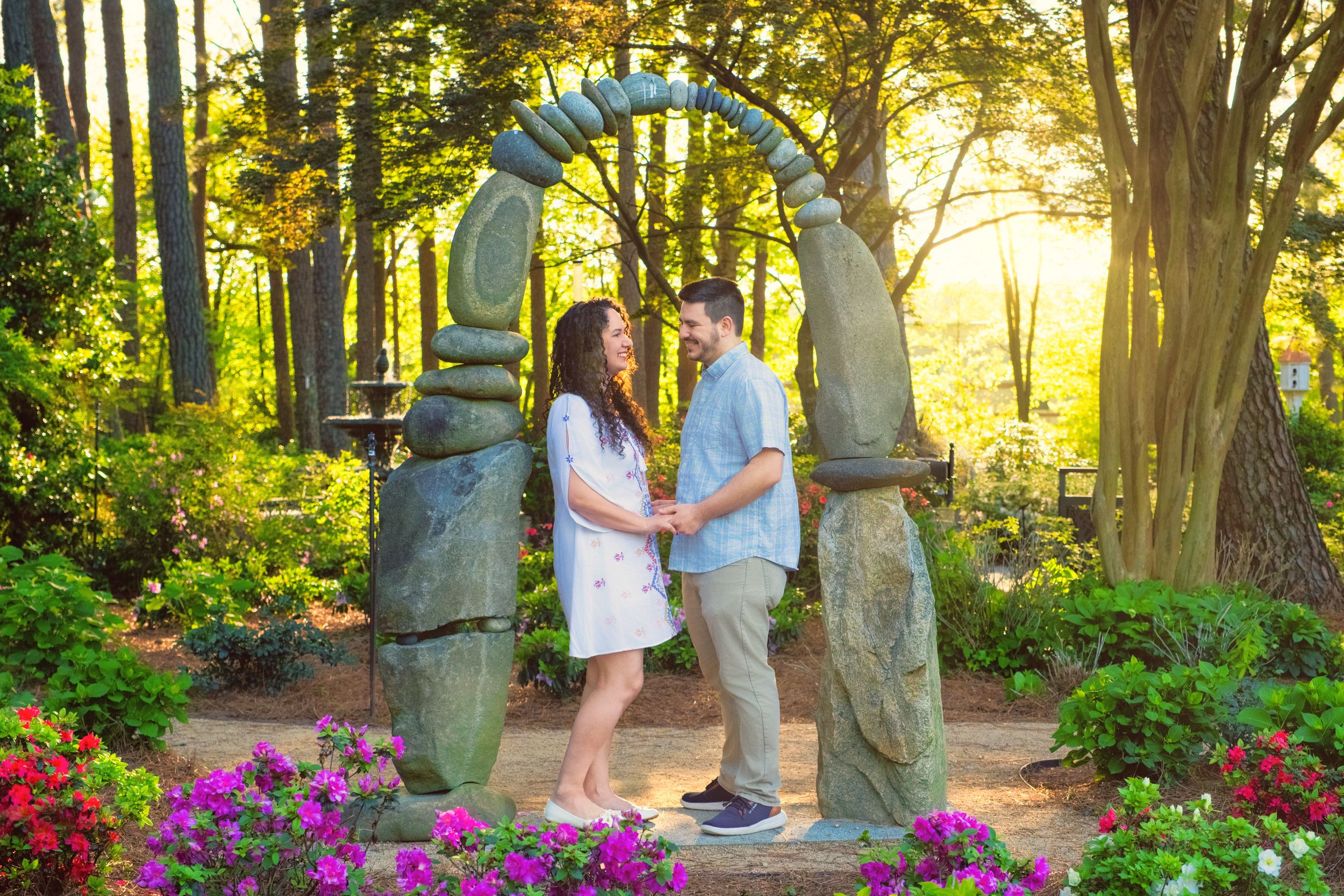 Couple embracing under a stone archway in a lush garden with colorful flowers and glowing sunset.