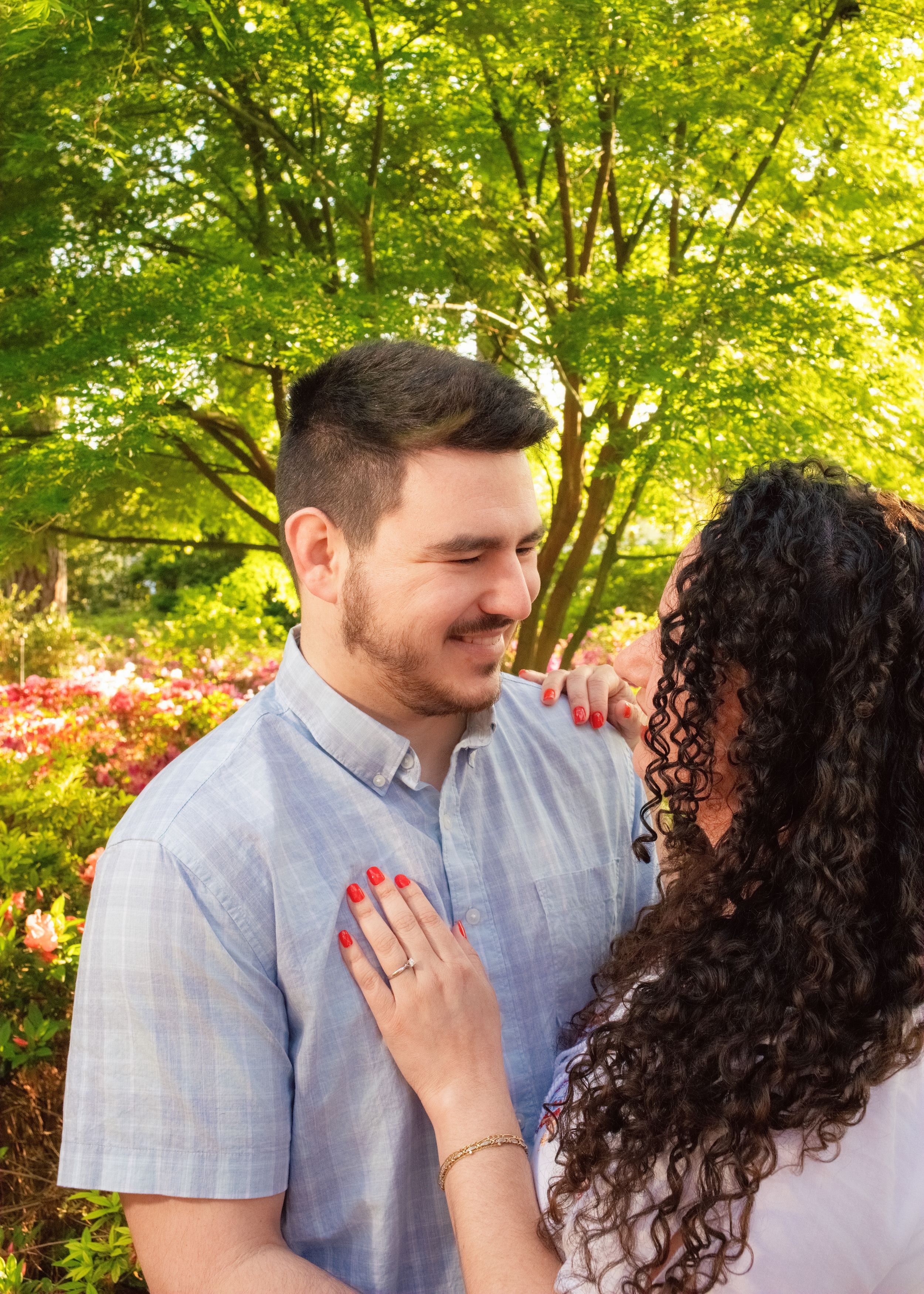 A couple shares an intimate moment in a garden with blooming flowers and lush green trees in the background.